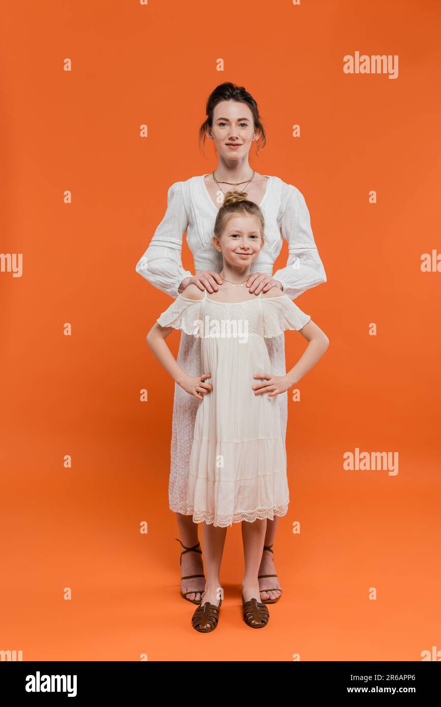 Moderne Erziehung, Sommerkleidung, Frau umarmt ihre Tochter und steht zusammen in weißen Sonnenkleidern auf orangefarbenem Hintergrund, weibliche Bindung, pfiffig Stockfoto