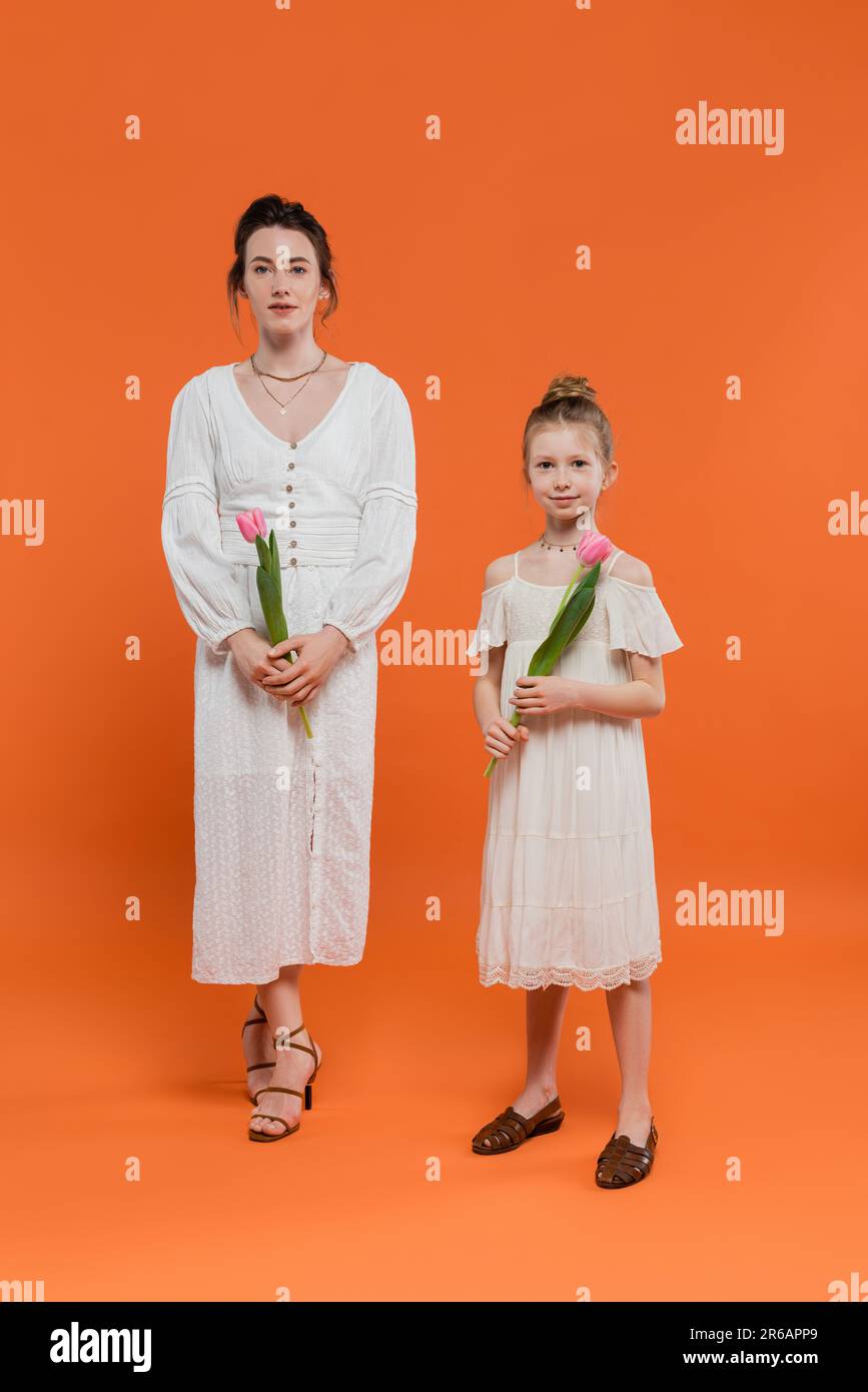 Glückliche Mutter und Tochter mit Tulpen, junge Frau und Mädchen mit Blumen und auf orangefarbenem Hintergrund, Familienstil, fröhlicher Anlass, Mode Stockfoto