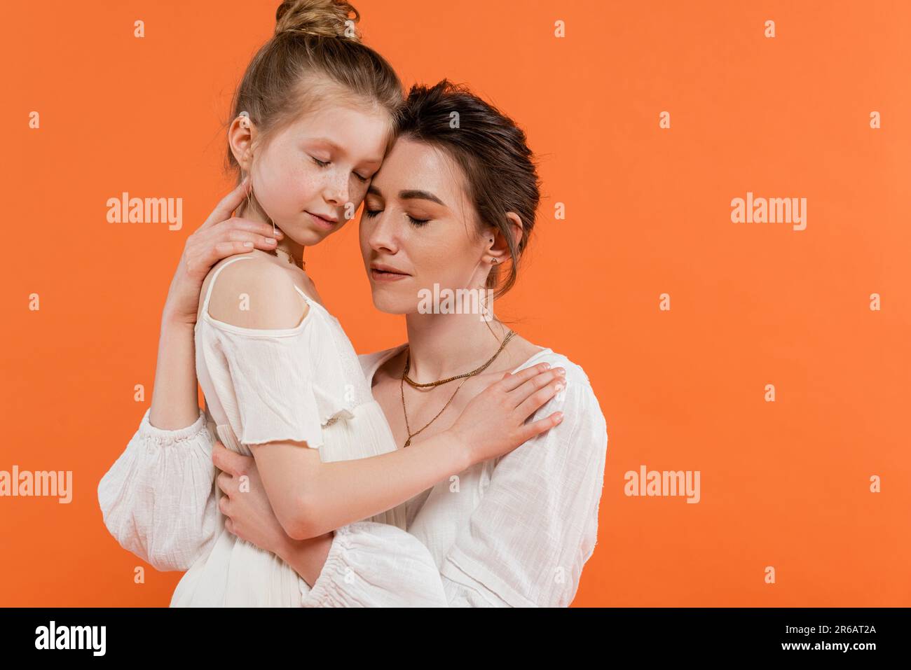 Mütterliche Liebe, Mutter und Tochter umarmen sich auf orangefarbenem Hintergrund geschlossene Augen, weiße Sonnenkleider, weibliche Bindung, moderne Erziehung, Liebe und Stockfoto