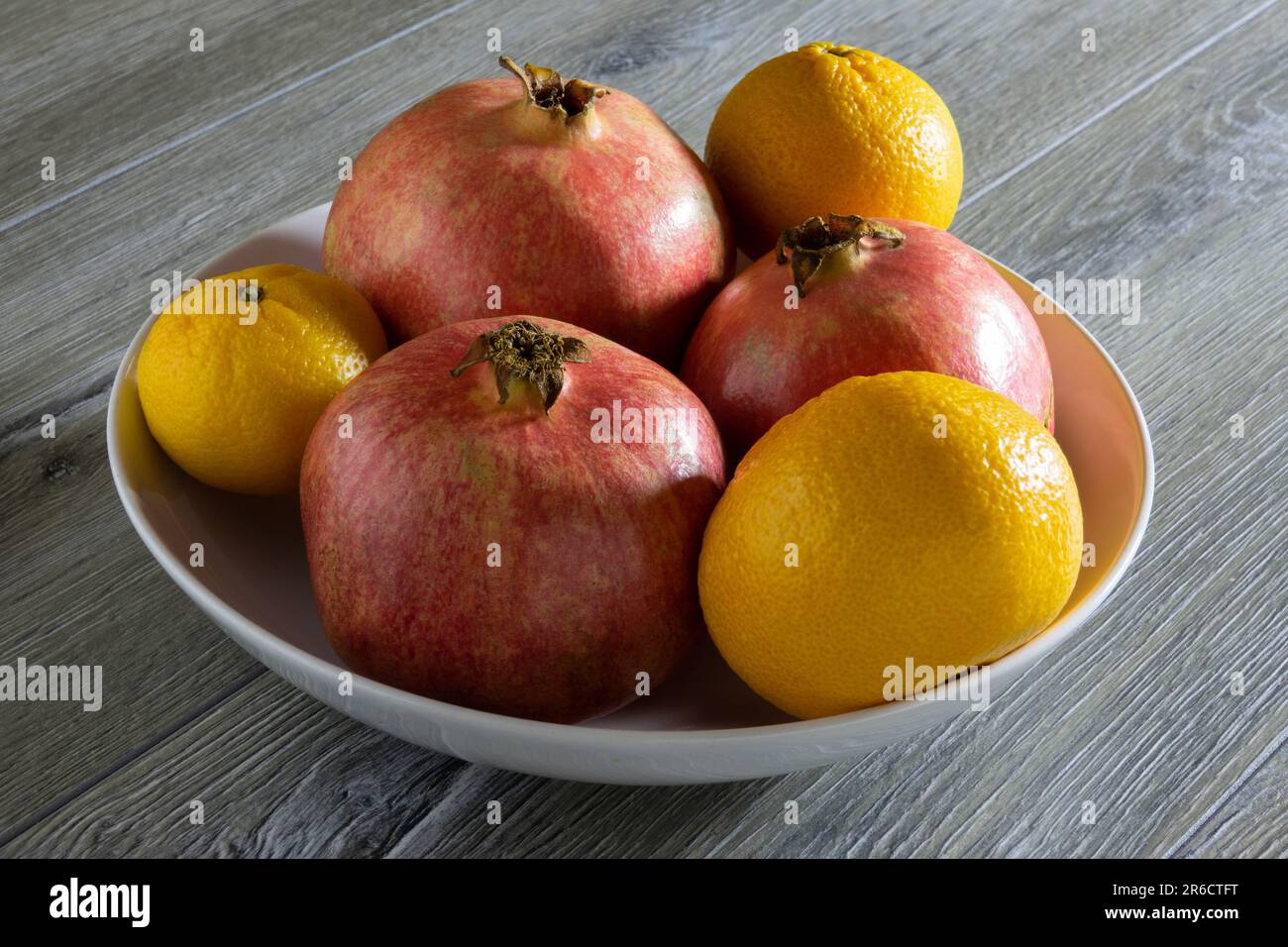 Drei frische, reife Granatäpfel und drei Orangen auf einem Teller auf einem grauen Holztisch. Nahaufnahme Stockfoto