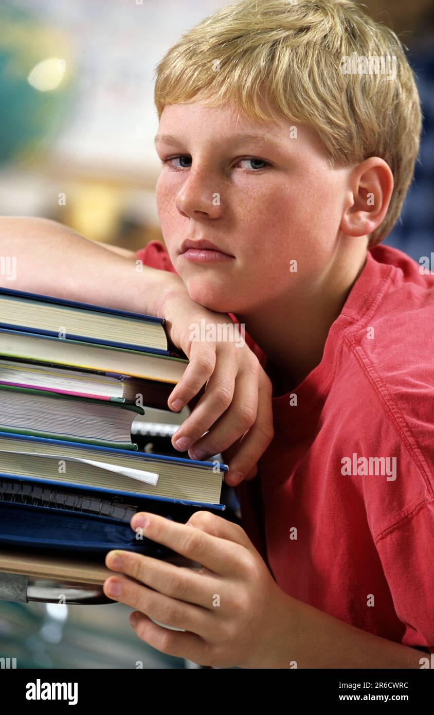 Porträt eines abgenutzten Jungen in der Schule, der im Unterricht nicht aufpasst, sich auf einen großen Stapel Bücher lehnt Stockfoto