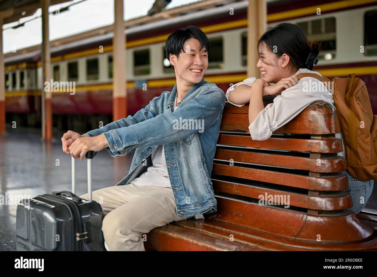 Ein hübsches, junges asiatisches Touristenpaar mit ihren Koffern spricht gerne auf einer Bank auf dem Bahnsteig, bevor es gemeinsam in den Zug steigt. Flitterwochen, Stockfoto