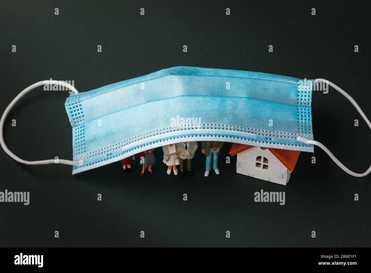Konzept zum Thema Hausquarantäne, Plastikspielzeugleute und ein Haus unter einer medizinischen Einwegmaske auf dunklem Hintergrund Stockfoto