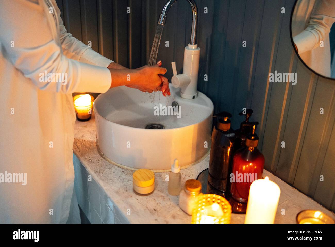Ein junges Mädchen in weißen Kleidern wäscht sich die Hände unter fließendem Wasser über dem Waschbecken. Kosmetik und Reinigungsmittel, angezündete Duftkerzen auf einem Marmortisch. Stockfoto