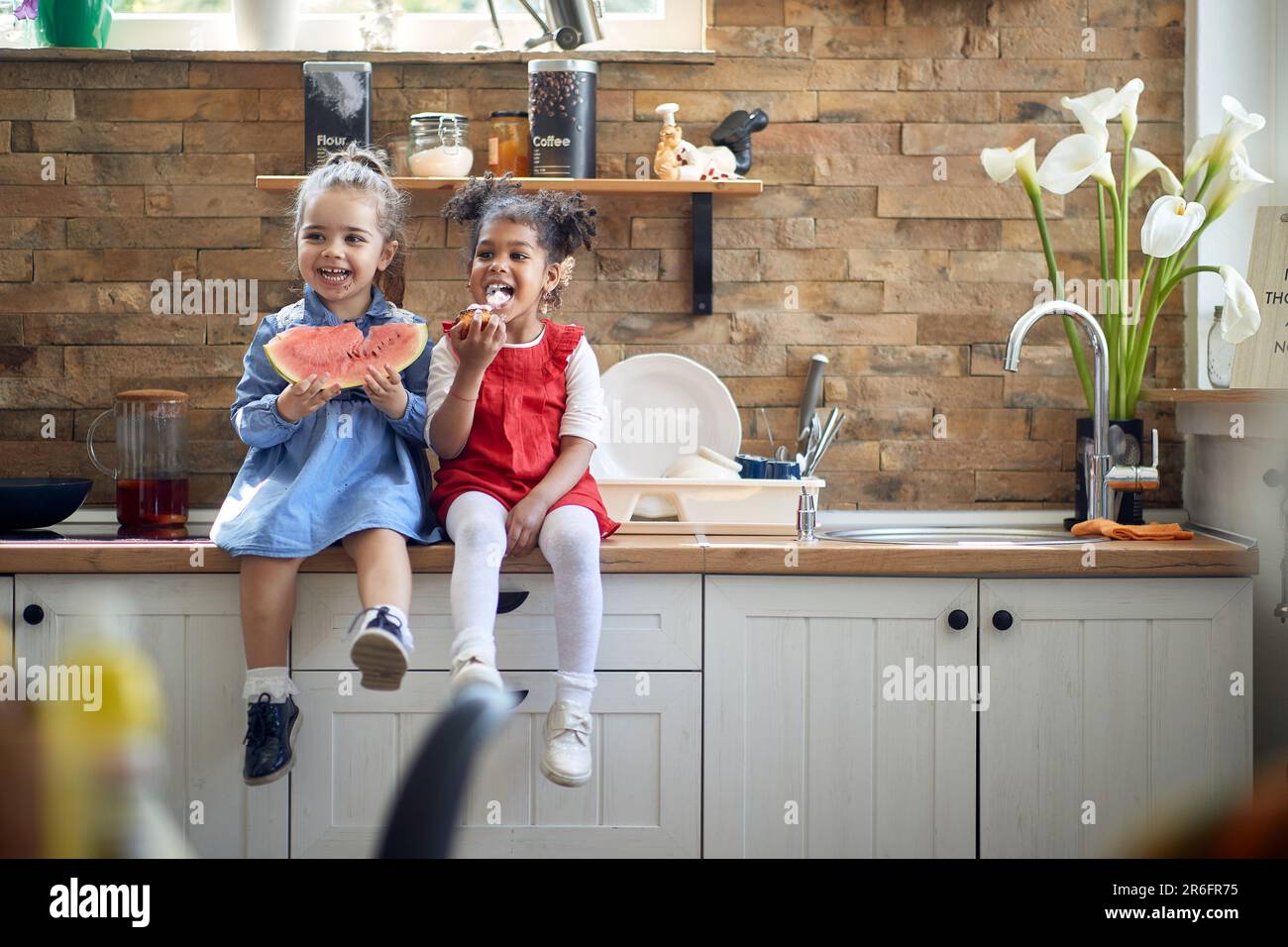 Zwei süße kleine Mädchen, die auf einer Kithcne-Arbeitsfläche sitzen und sich albern benehmen und einen gesunden Snack zusammen genießen. Zuhause, Familie, Lifestyle-Konzept. Stockfoto