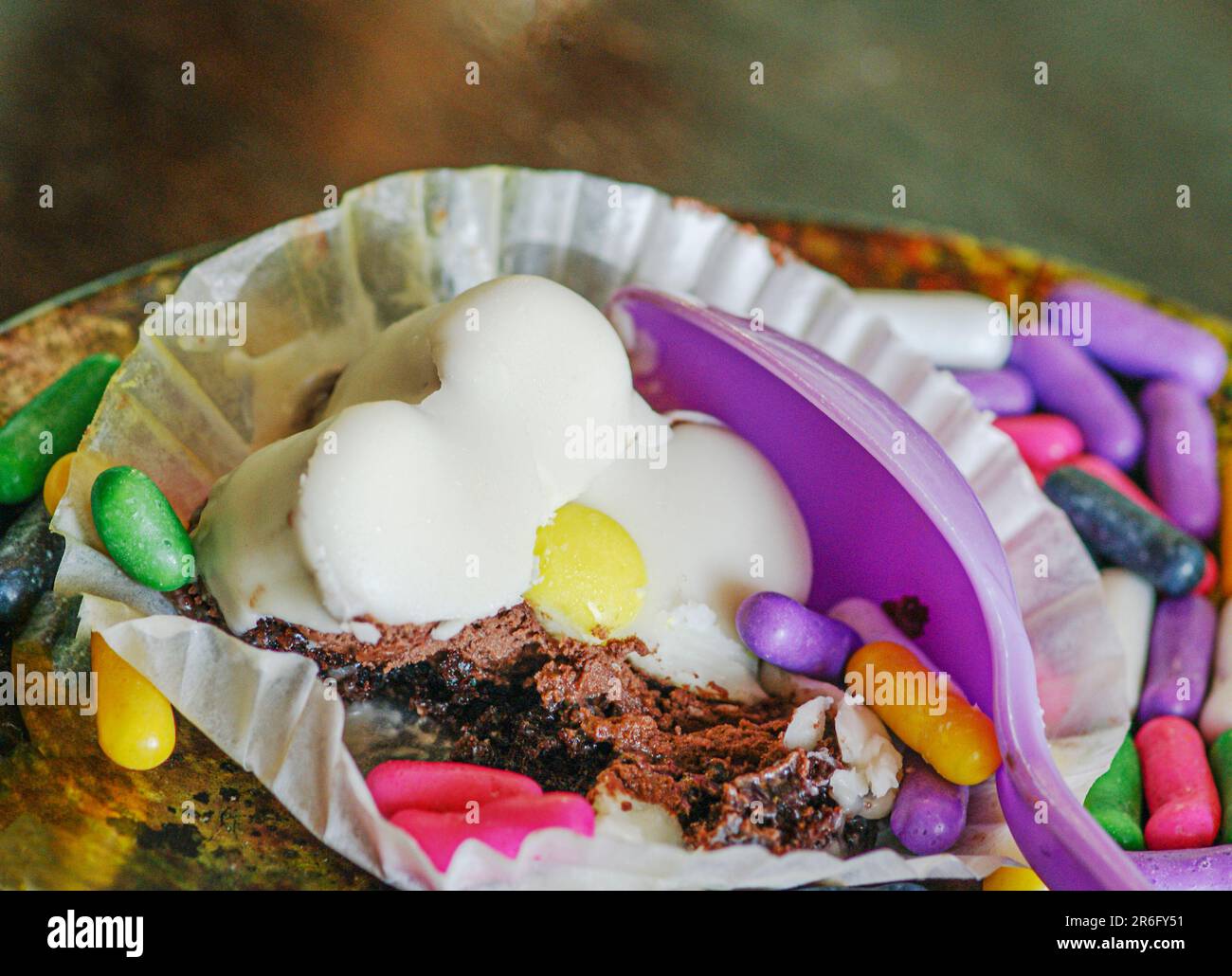 Ein teilweise aufgegessen Dessert ist in einem Cupcake-Halter mit bunten Süßigkeiten. Das Dessert besteht aus Schokoladenkuchen-Pudding mit lila Plastiklöffel. Stockfoto