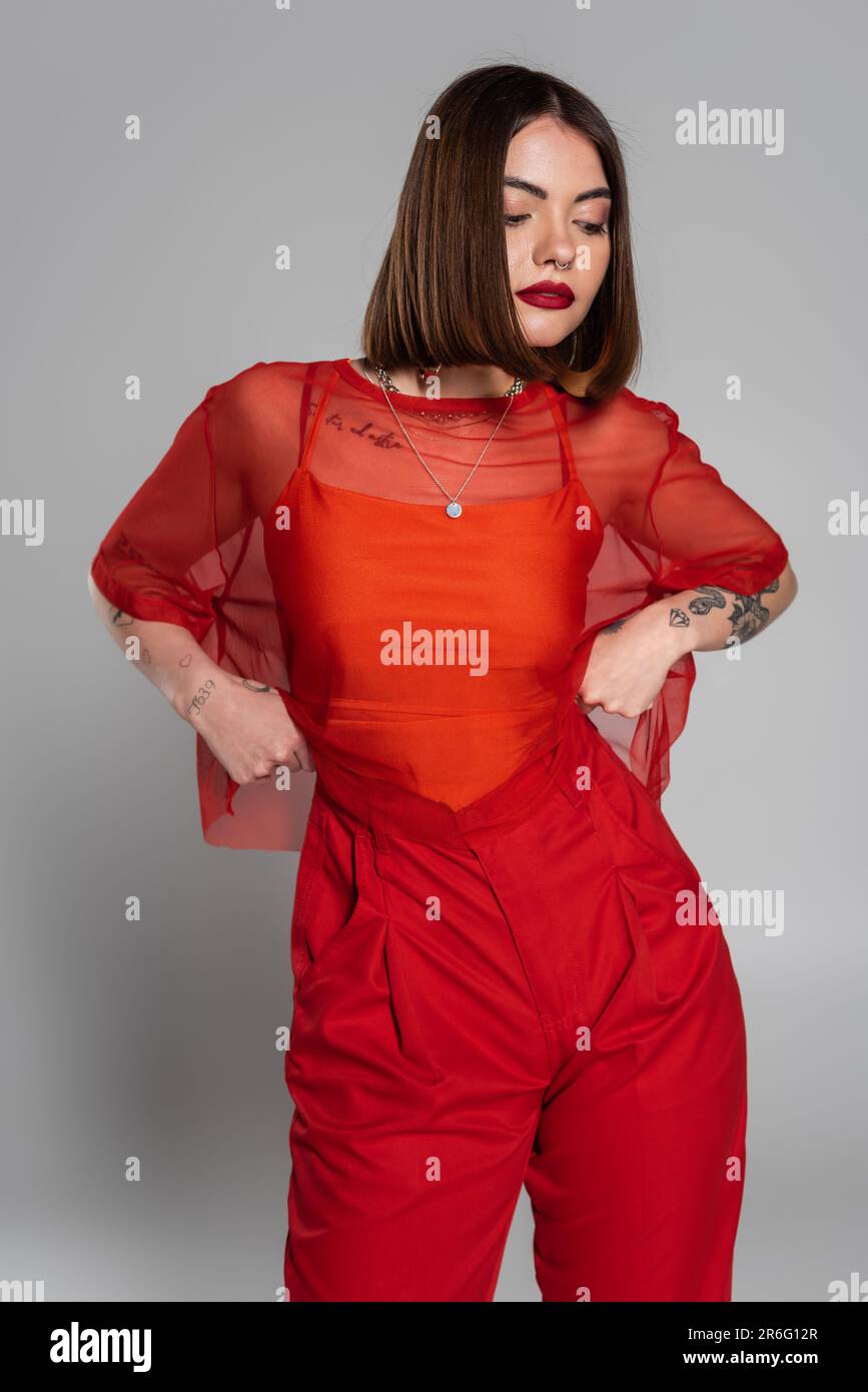 Stylisches rotes Outfit, tätowierte und brünette Frau mit kurzen Haaren und Nasenlöchern in transparenter Bluse und Hose auf grauem Hintergrund, moderne Stockfoto
