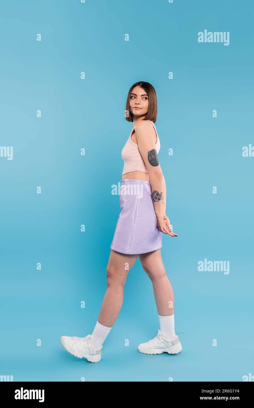 Modetrend, brünette junge Frau mit kurzen Haaren im Tanktop, Rock und weiße Turnschuhe auf blauem Hintergrund, Freizeitkleidung, gen z Mode, pe Stockfoto