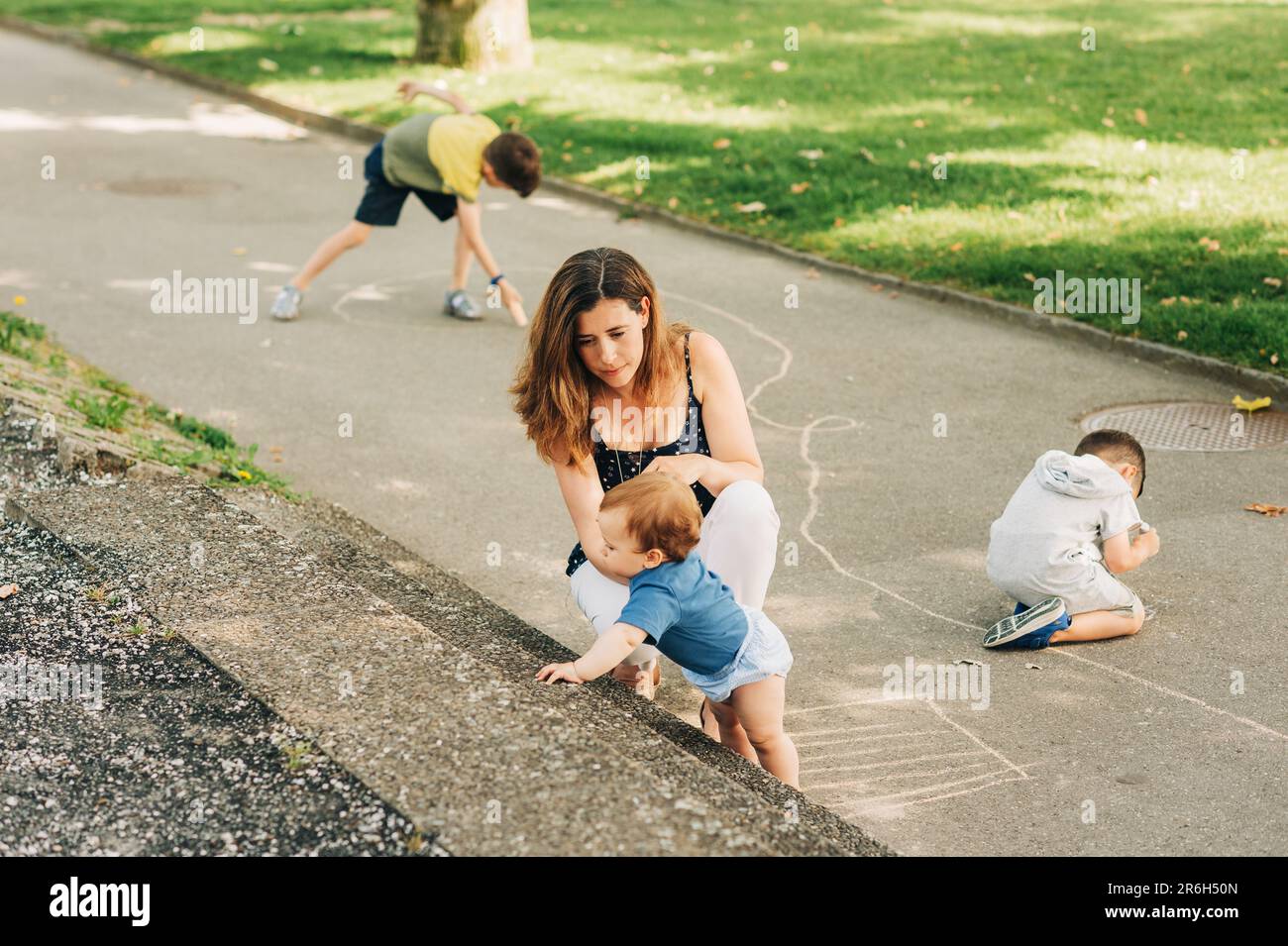 Glückliche Mutter, die Zeit mit Kindern im Sommerpark verbringt, mit dem Baby spielt, Jungs mit Kreide malt Stockfoto