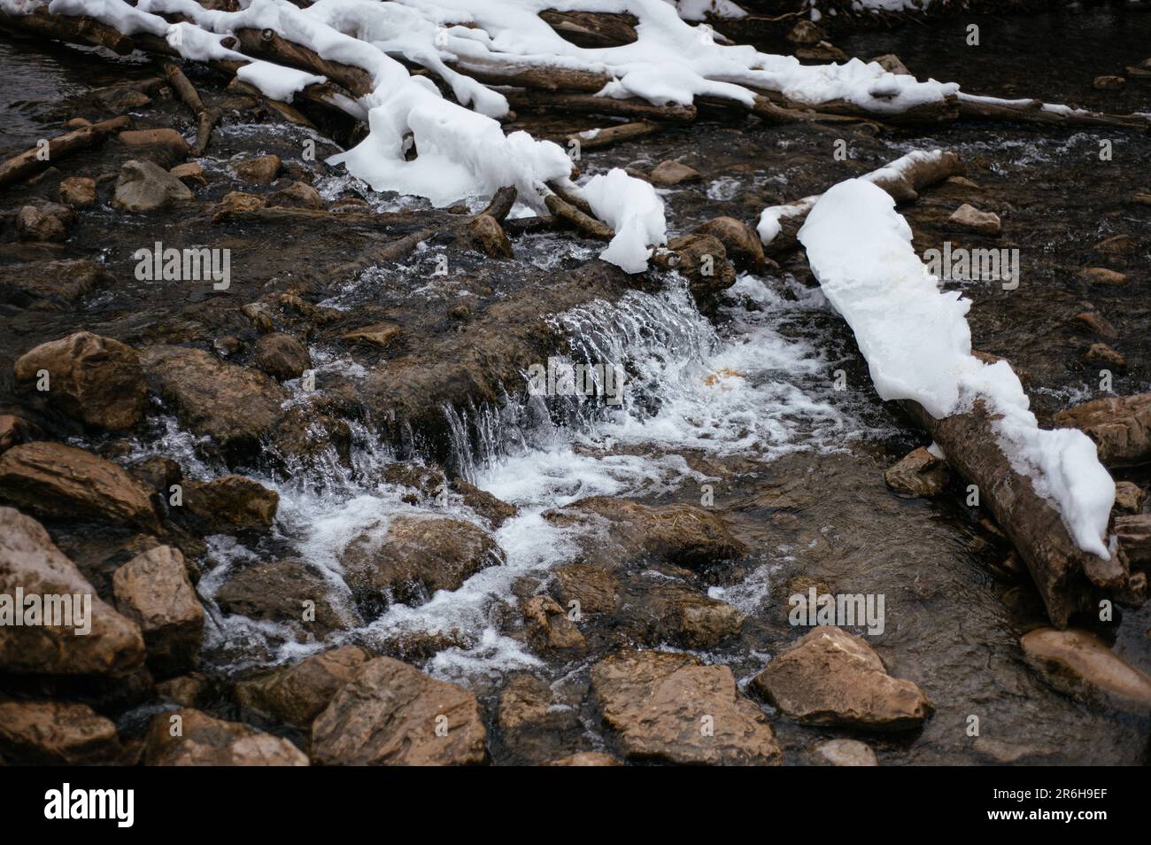 Eine idyllische Winterszene mit einem ruhigen Bach mit einer Decke aus Schnee, Eis und kleinen Steinen, die im Sonnenlicht glitzern Stockfoto Eine idyllische Winterszene mit einem ruhigen Bach mit einer Decke aus Schnee, Eis und kleinen Steinen, die im Sonnenlicht glitzern Stockfoto