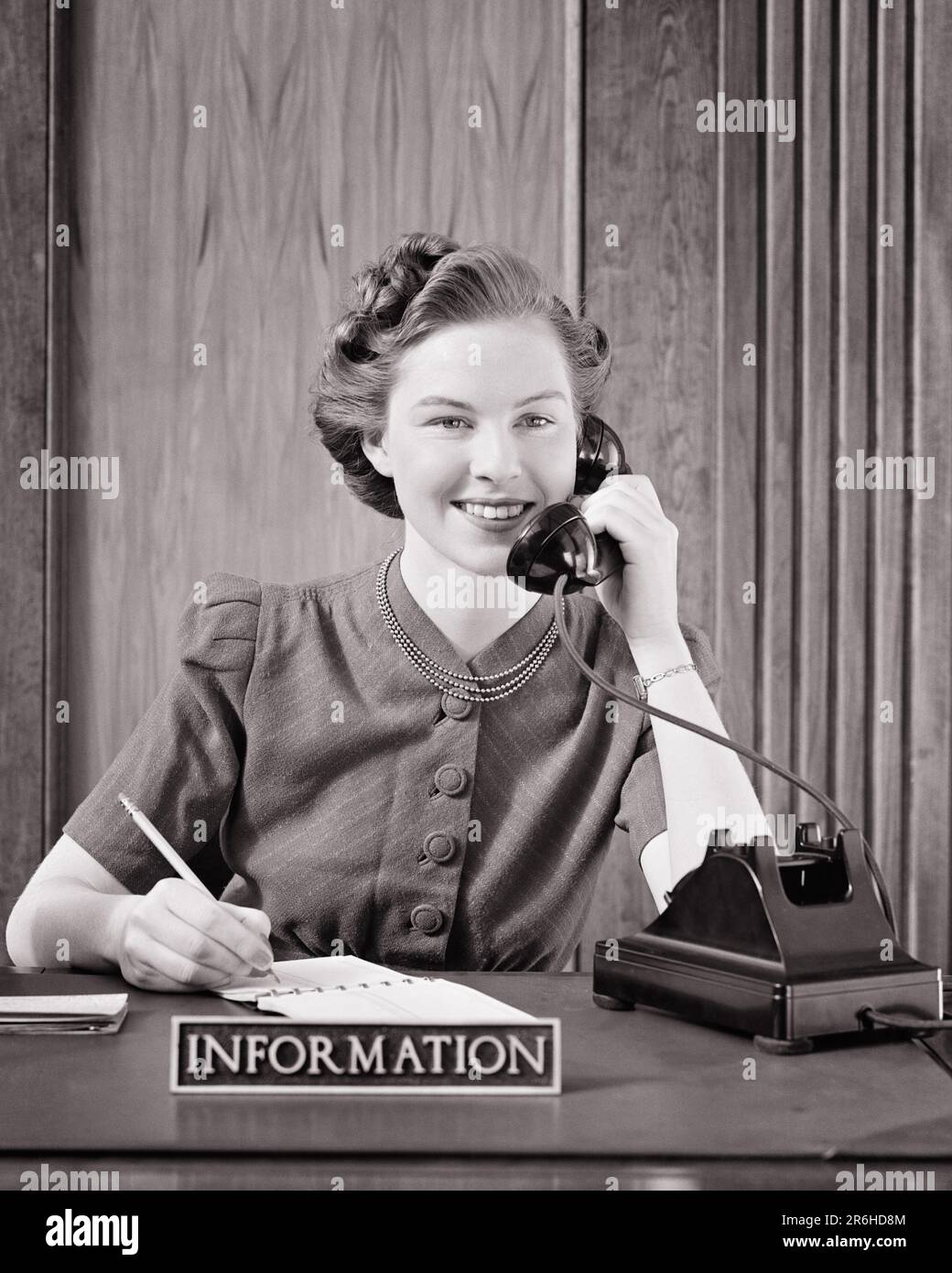 1940S LÄCHELNDE SEKRETÄRIN AN DER REZEPTION, DIE AM INFORMATIONSSCHALTER SITZT UND TELEFONIERT, SICH NOTIZEN MACHT UND IN DIE KAMERA SCHAUT - O522 HAR001 HARS EMPFANGSMITARBEITER WILLKOMMEN KOMMUNIKATION KARRIERE JUNGER ERWACHSENER NOTIZEN INFORMATIONSASSISTENT ZUFRIEDEN LEBENSSTIL ZUFRIEDENHEIT ANGESTELLTE WEIBLICHE JOBS STUDIO SHOT GESUNDHEIT KOMMUNIZIERT KOPIE WELTRAUM FREUNDSCHAFT FRAUEN BERUF VERTRAUEN B&W AUGENKONTAKT FÄHIGKEIT BERUF GLÜCK FÄHIGKEITEN KOPF UND SCHULTERN FRÖHLICH KUNDENSERVICE KARRIERE FRISUR BÜROANGESTELLTER BEI ON OCCUPATIONS TELEFONE LÄCHELN VERBINDUNG GAL FRIDAY TELEFONE ADMINISTRATOR JOYFUL Stockfoto