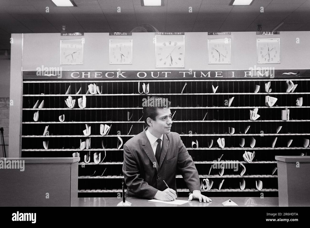 1960S HOTELANGESTELLTER AN DER REZEPTION, NACHRICHTENFENSTER, CHECKOUT BIS 1 UHR, VERSCHIEDENE WELTZEITEN, EMPIRE HOTEL NEU YORK CITY USA - Q68251 CPC001 HARS-ANGESTELLTE JOBS USA COPY SPACE PERSONEN USA VON AMERIKA MÄNNER BERUF MANAGEMENT EMPIRE CONFIDENCE B&W NORDAMERIKA QUALIFIKATIONSANZUG UND KRAWATTE BESCHÄFTIGUNG FÄHIGKEITEN KUNDENSERVICE EMPFANG KARRIERE WISSEN WIRD ERFASST DIE CLERKS AUTHORITY NYC OCCUPATIONS NEW YORK CITIES UNTERSTÜTZEN VERSCHIEDENE NEW YORK CITY SLOTS PM JUNGER ERWACHSENER IN SCHWARZ UND WEISS DIE BOTSCHAFT DER KAUKASISCHEN ETHNISCHEN HERKUNFT IST ALTMODISCH Stockfoto