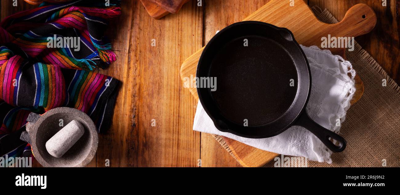 Leere gusseiserne Pfanne auf dem Schneidebrett. Kochhintergrund, farbenfroher traditioneller mexikanischer Stoff und kleine Molkajete auf rustikalem Holztisch. Panoram Stockfoto