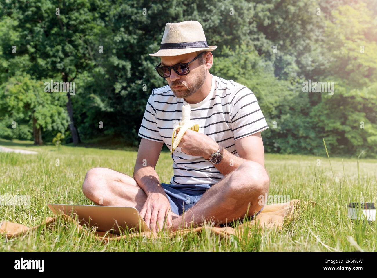 Ein Mann isst Banane, während er auf einem grünen Rasen im Park sitzt und am Laptop arbeitet. Stockfoto