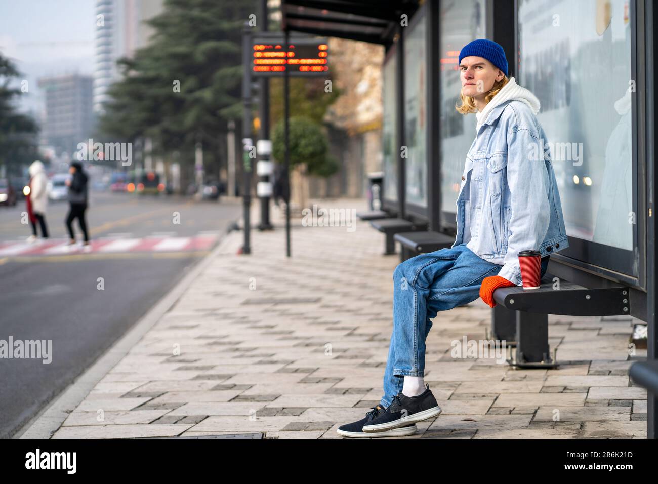 Der junge blonde Student wartete sehr lange auf den Transport am Morgen. Ein Mann an der Bushaltestelle mit Kaffee weg Stockfoto