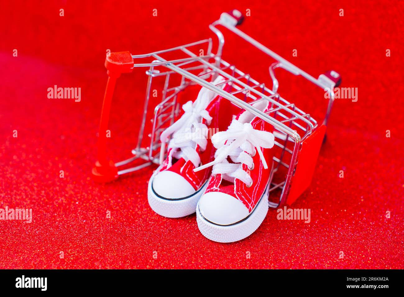 Ein Paar rote Mini-Segelschuhe in einem umgedrehten Miniaturwagen mit glitzerndem rotem Hintergrund. Einkaufen, Geschenke machen, Urlaub machen Stockfoto