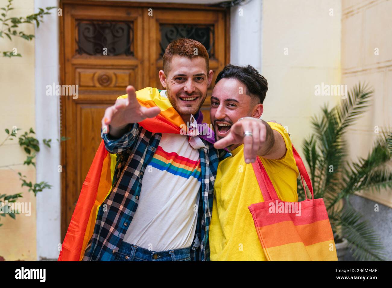 Fröhliche schwule Freunde umarmen sich, zeigen stolz von der Tür nach außen, geschmückt mit LGBT Pride Accessoires Stockfoto