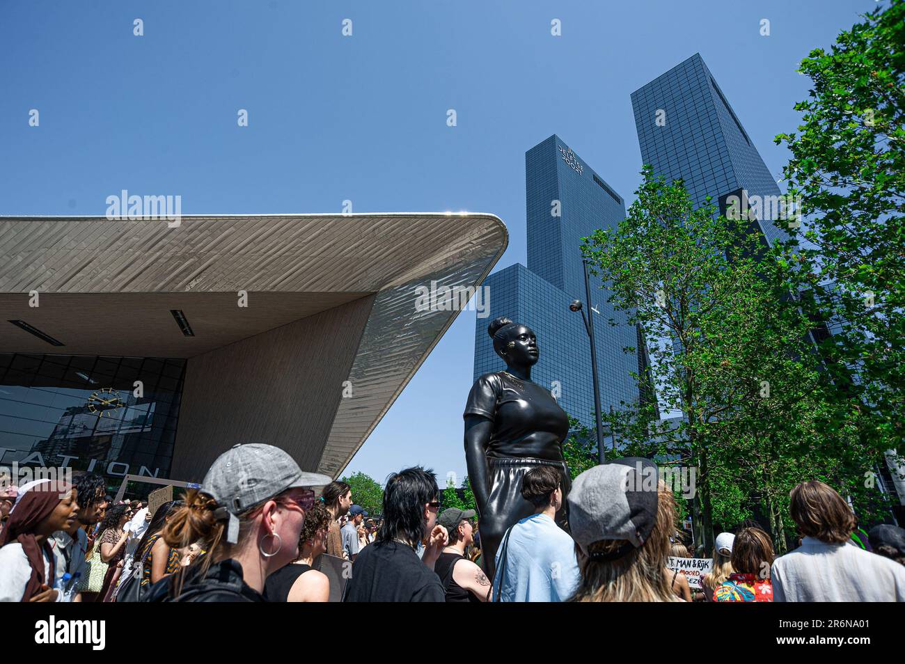 Demonstranten durch die Statue „Momente Containment“, während des Protests „Black Lives Matter“. Black Live Matter Protest für die belgische Studentin Sanda Dia by Moments Convenient, ein vier Meter hohes Statut einer schwarzen Frau in Jogginghosen und Turnschuhen mit Blick auf Rotterdams Stationsplein, das Statut wird vom britischen Künstler Thomas J Price erschaffen. Black Lives Matter und andere, versammelt außerhalb von Rotterdams geschäftigem Stationsplein, für Sanda Dia, eine zwanzig Jahre alte belgische Farbstudentin, die seinen Tod bei einer Verbindungsinitiationszeremonie namens Reuzegom im Jahr 2018. Die Zeremonie war gleichbedeutend Stockfoto