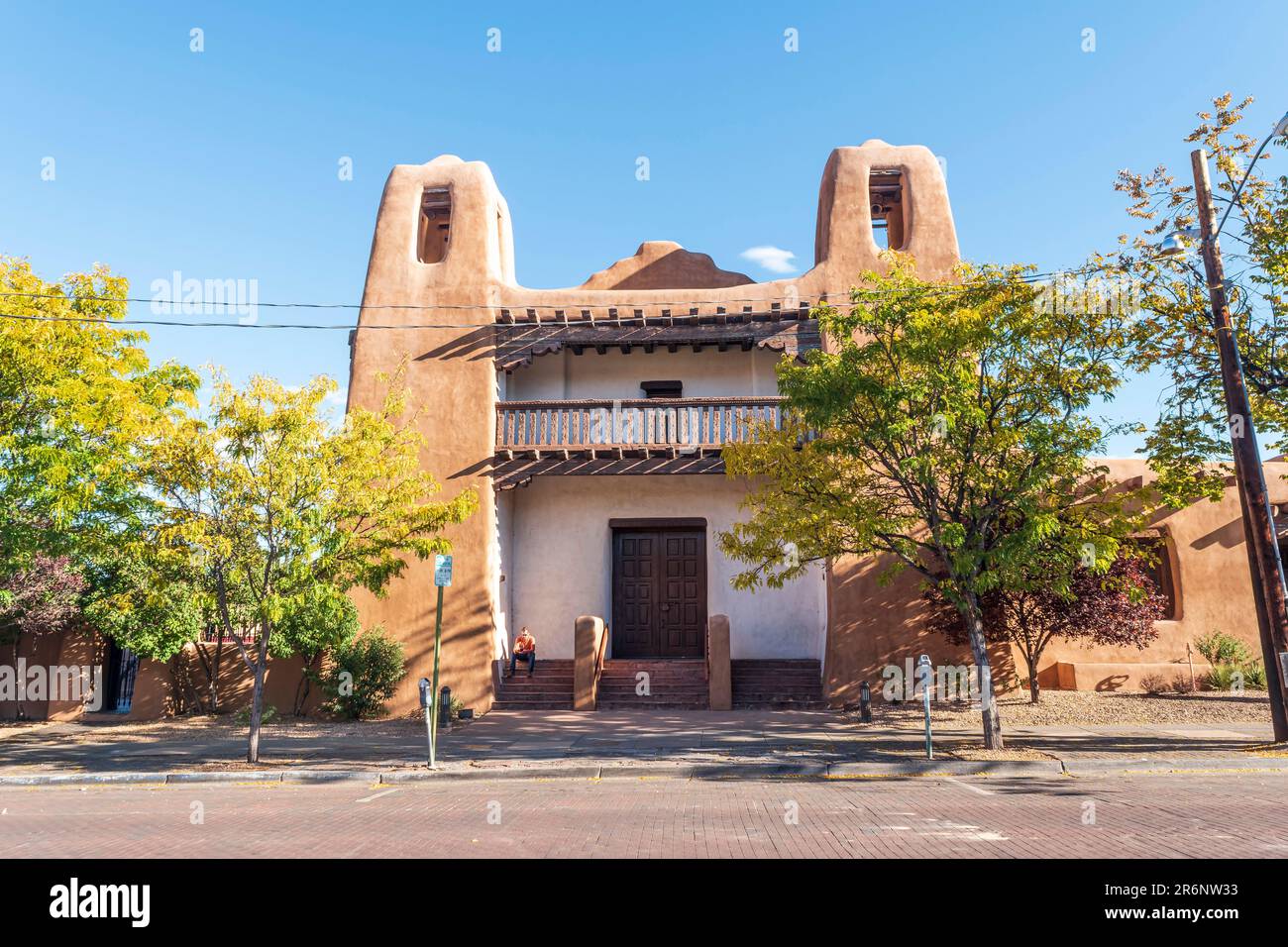 Horizontales Bild eines Mannes in einem orangefarbenen Hemd, der auf den Stufen des Gebäudes im Pueblo-Revival-Stil sitzt, in dem das New Mexico Museum of Art in San untergebracht ist Stockfoto