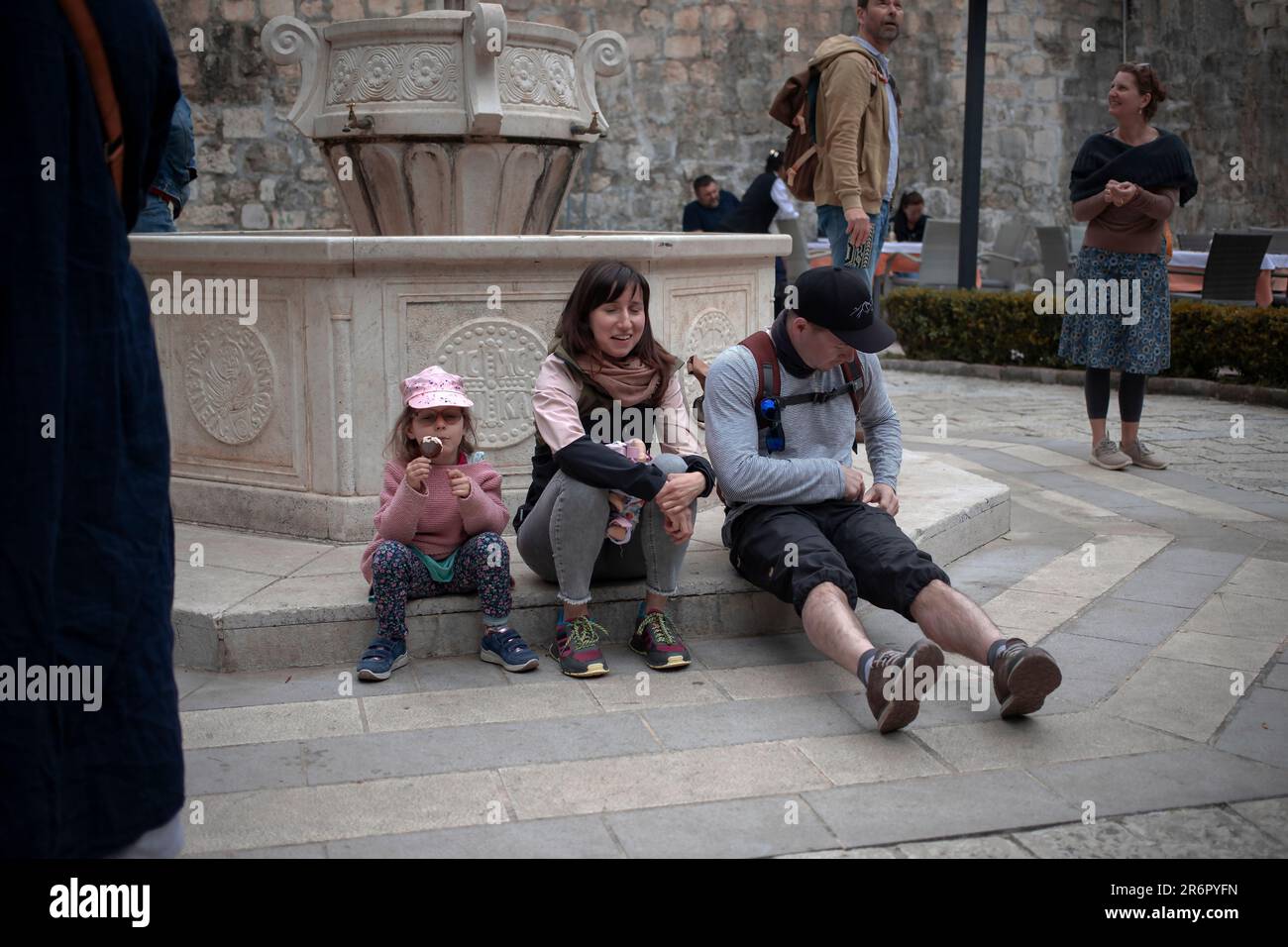 Kotor, Montenegro, 13. April 2023: Ein touristisches Paar mit einer Tochter, das eine Pause macht und auf dem Platz neben einem Trinkwasserbrunnen sitzt Stockfoto