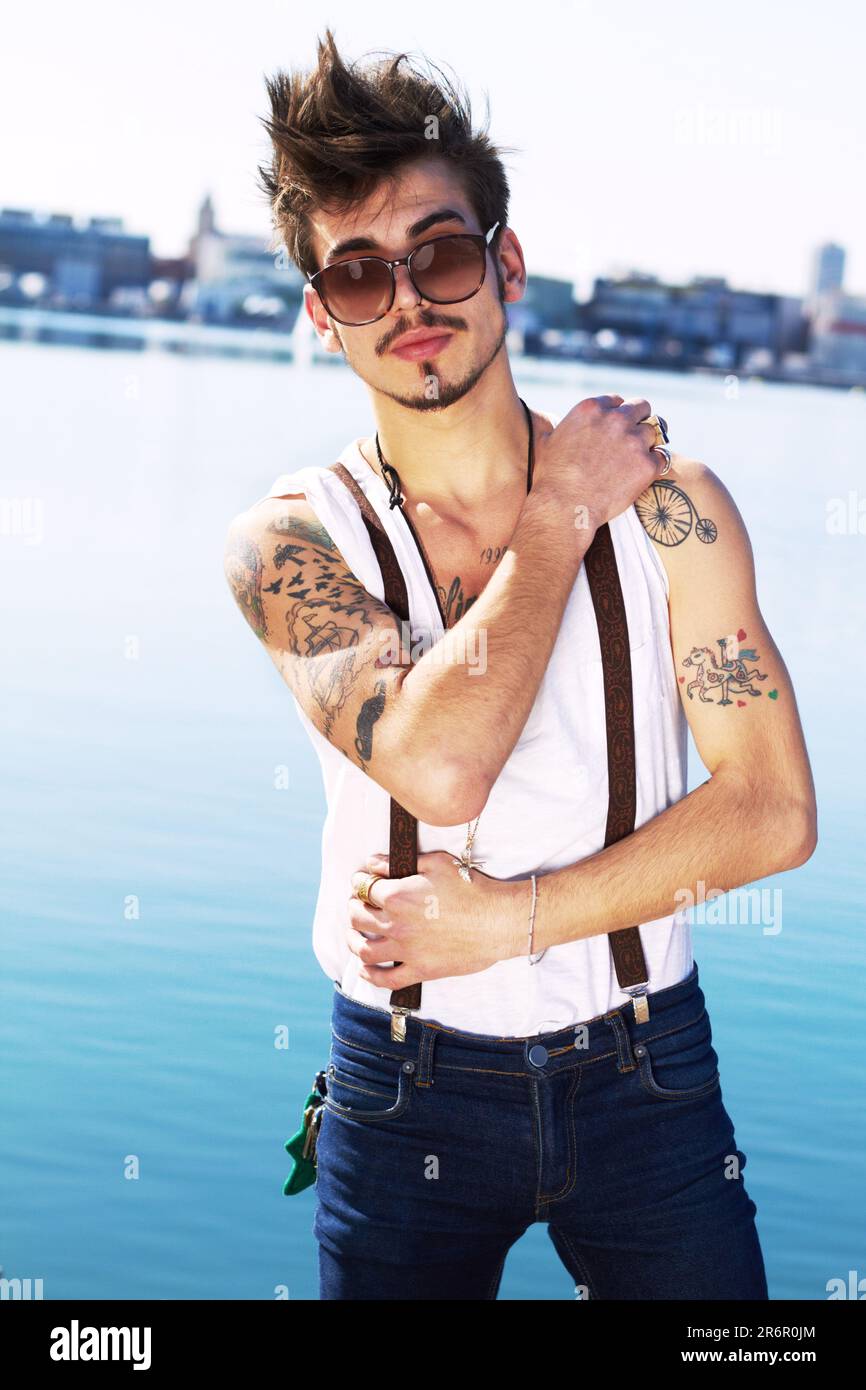 Sonnenbrille, Porträt und ein echter Mann auf dem Meer mit stilvollem Tattoo, Körperkunst und Mode. Punk, Ozean und trendiger Mann, der draußen in Spanien steht Stockfoto