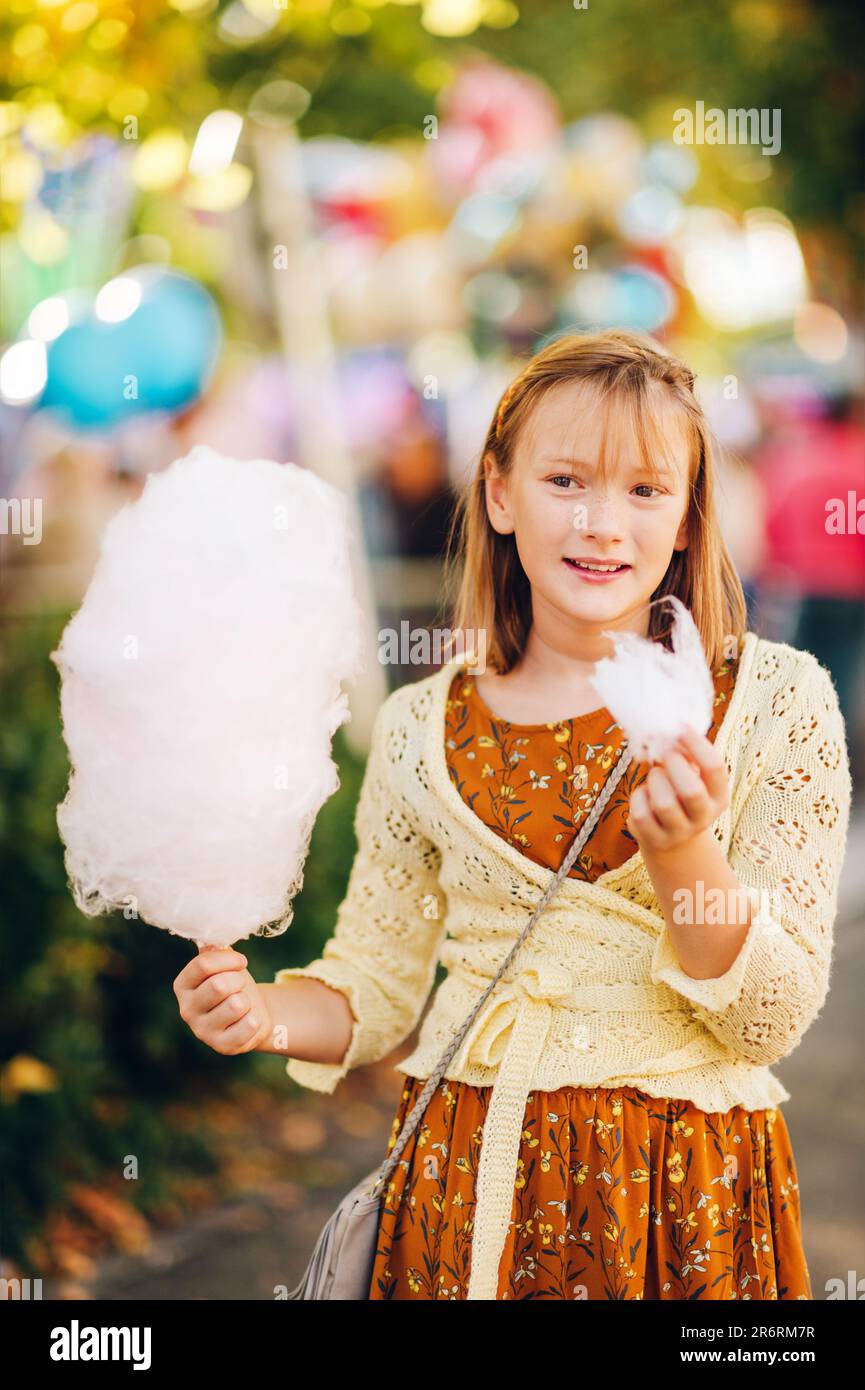 Hübsches junges Mädchen, das an einem sonnigen Tag auf dem Herbstkarneval Zuckerwatte isst, braunes Kleid und gelbe Strickjacke trägt Stockfoto