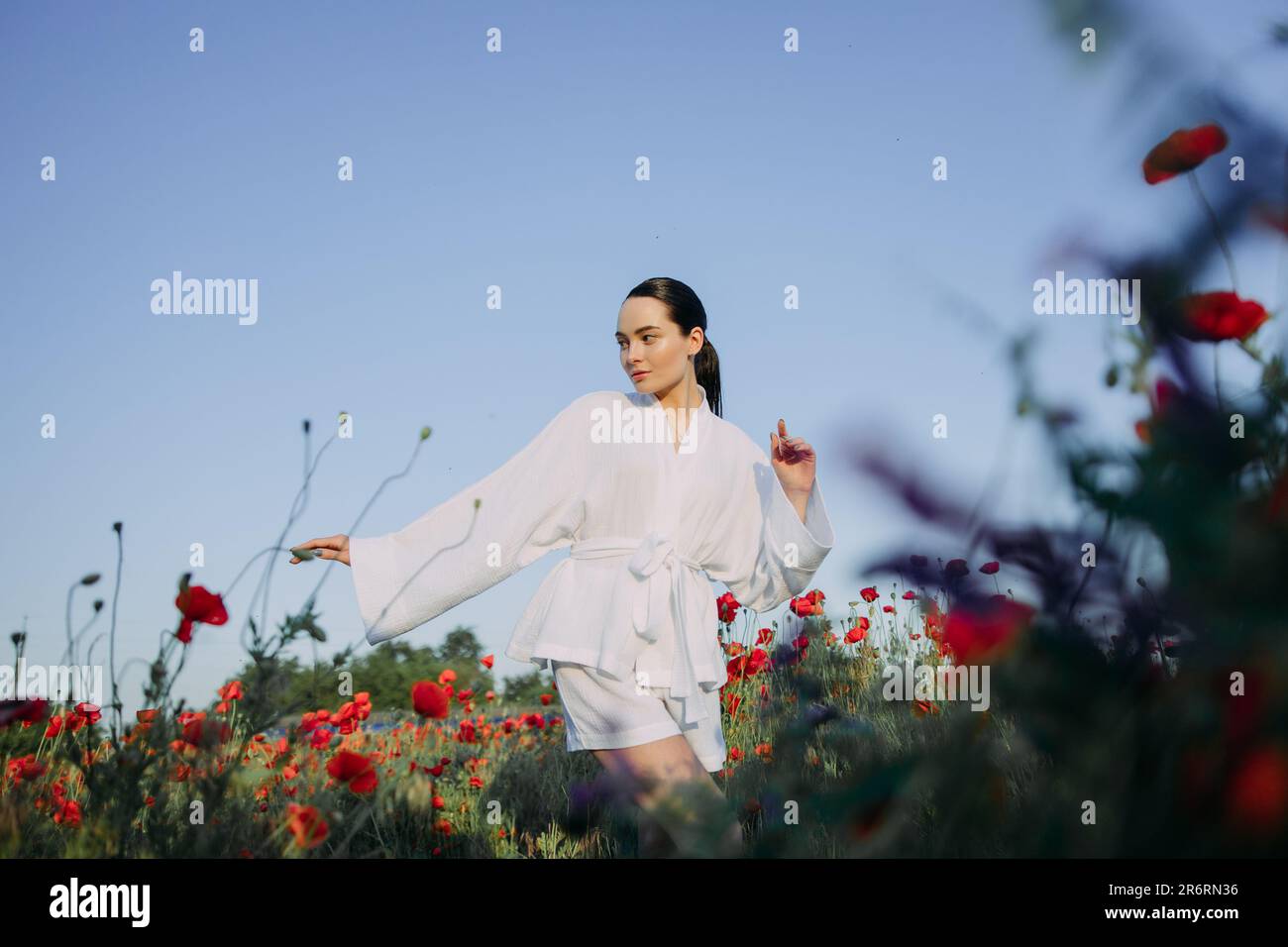 Junge Frau in weißer Kleidung, die auf blühenden Mohnwiesen vor dem Hintergrund des Himmels spazieren geht und sich amüsiert. Stockfoto