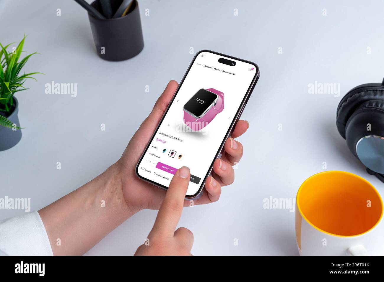 Smartwatch online mit Smartphone einkaufen. Person, die ein modernes Smartphone an einem Tisch im Haus hält, umgeben von einer Tasse Bleistifte, einer Zimmerpflanze und Stockfoto