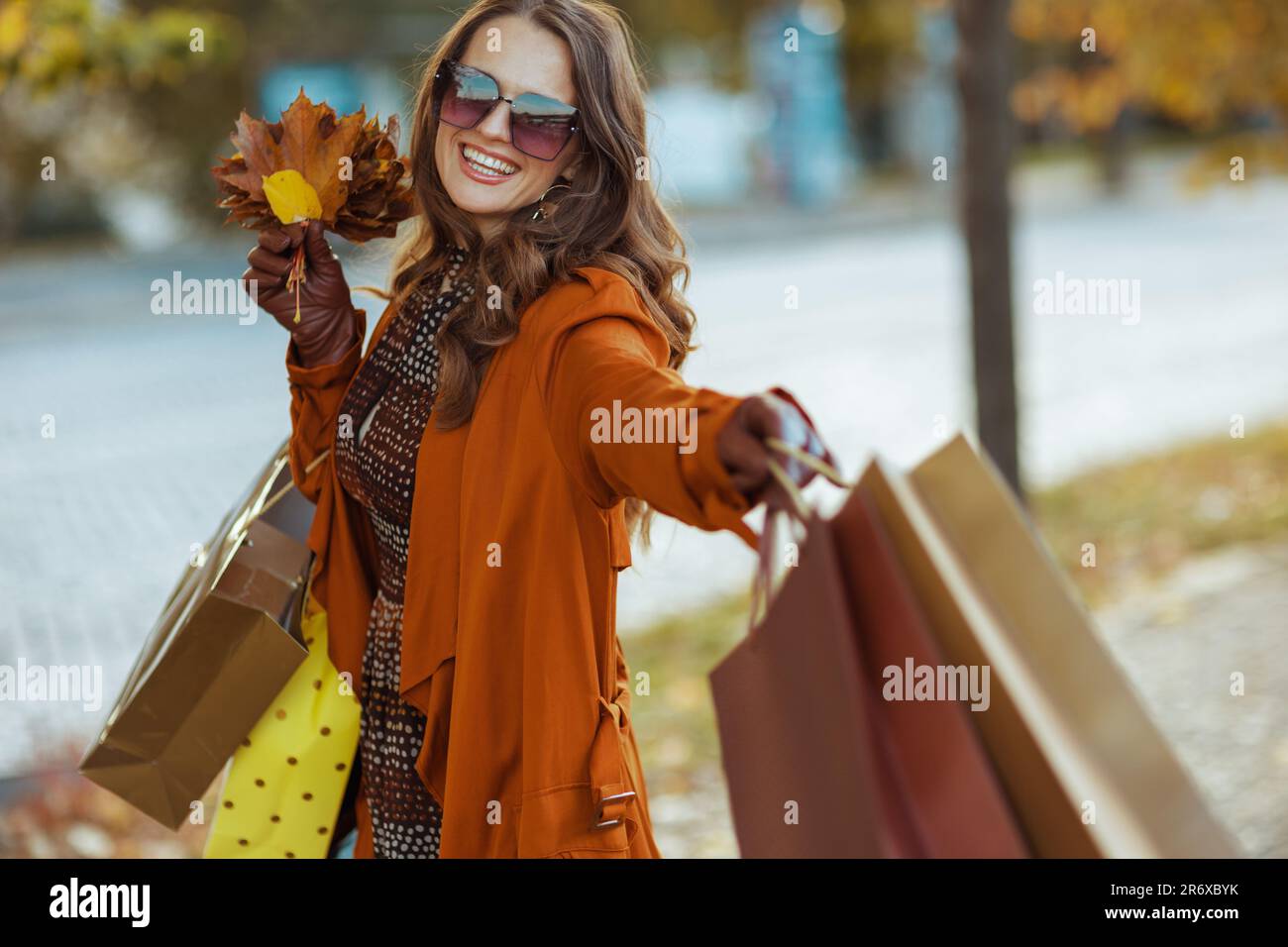 Hallo Herbst. Glückliche, stilvolle Frau in braunem Trenchcoat mit Einkaufstaschen und herbstgelben Blättern in der Stadt. Stockfoto