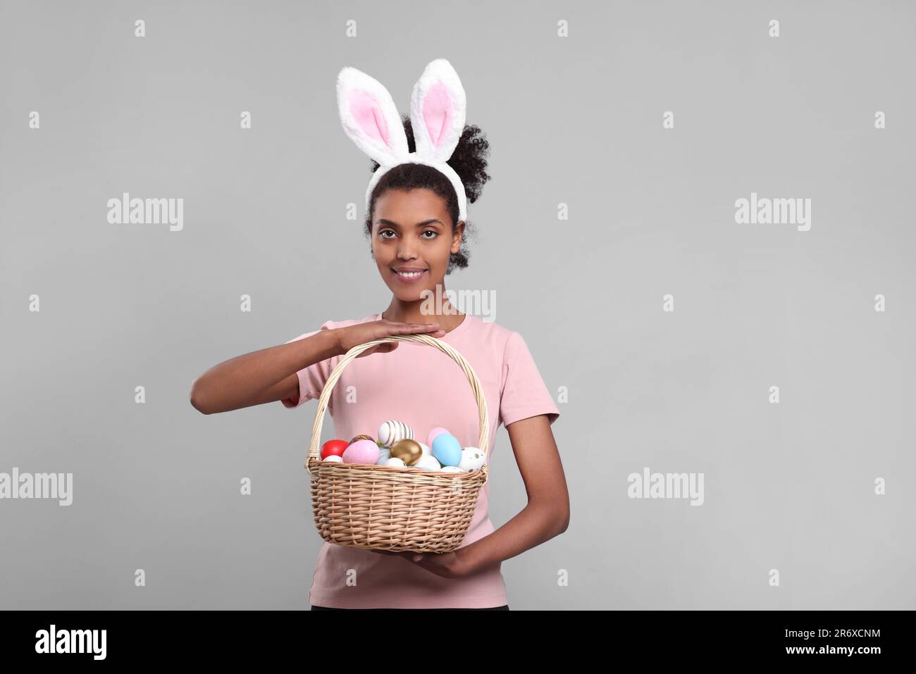 Glückliche afroamerikanische Frau mit Hasenohren Stirnband mit Korb aus Korb mit Ostereiern auf grauem Hintergrund Stockfoto