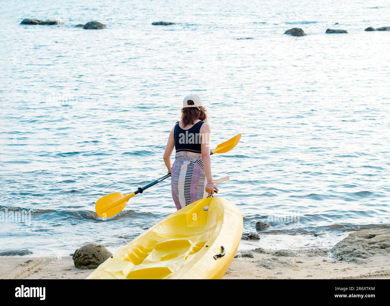 Asiatische Frau mit schwarzem Tanktop und Deckel, die ein gelbes Kajak schleppt und ein Paddel auf dem Sand zum Meer hält. Glückliche Frau mit lustiger Aktivität am Strand f Stockfoto