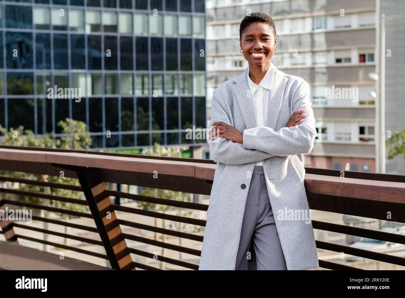 Fröhliche afrikanische Geschäftsfrau, die in die Kamera schaut. Sie kreuzt ihre Arme und hat Bürogebäude im Hintergrund Stockfoto