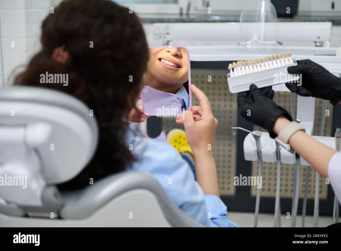 Rückfahrfrau im Zahnarztstuhl, lächelt Spiegel an, Zahnarzt benutzt Zahnschattierung, mit Vergleichsplatten Stockfoto
