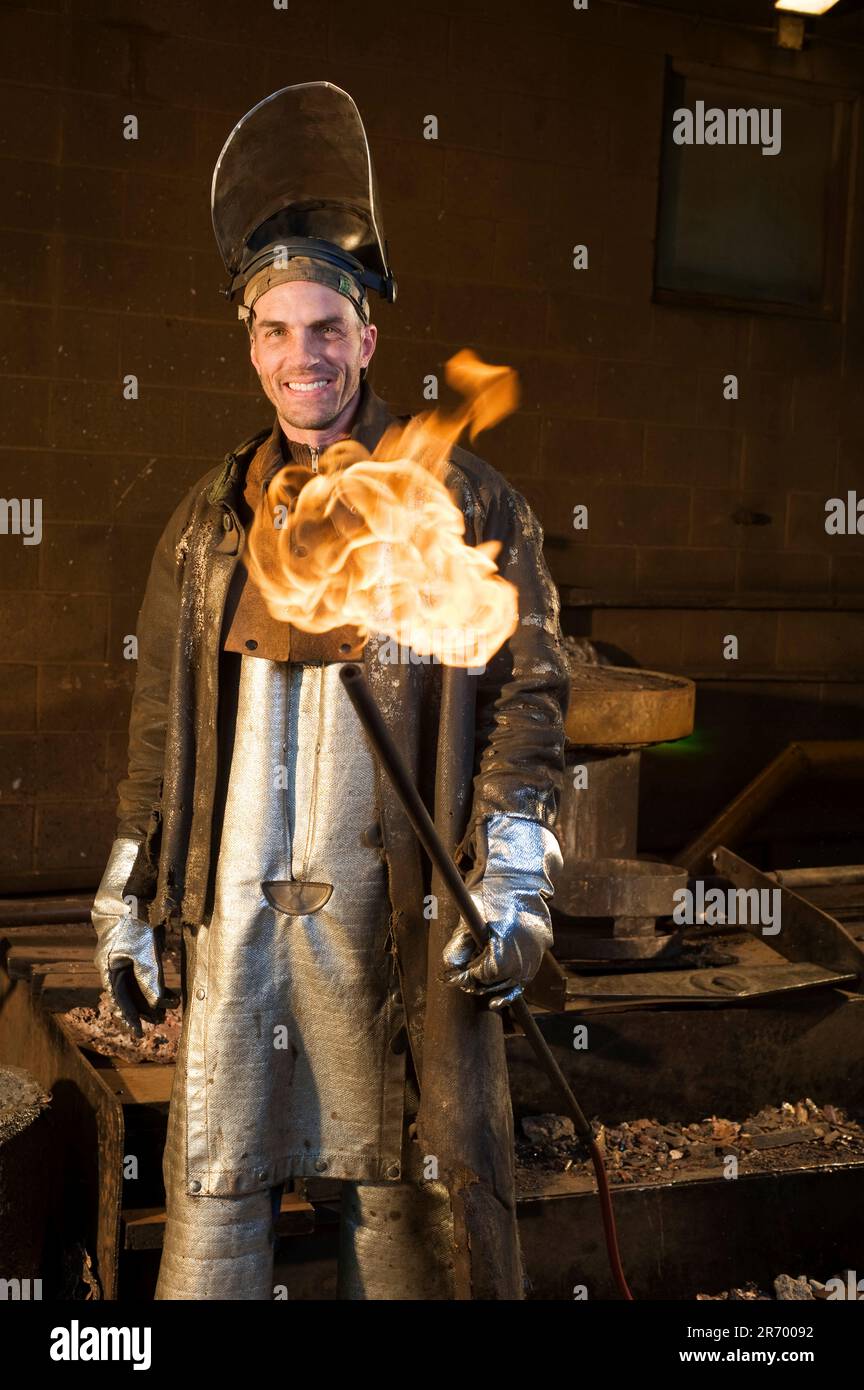 Ein Mann in einer Foudry Jacke, Overalls und einem Schutzhelm, mit hochgehobenem Gesichtsschutz, mit einer brennenden Röhre in einer Hand und einem Lächeln in der Kamera. Stockfoto