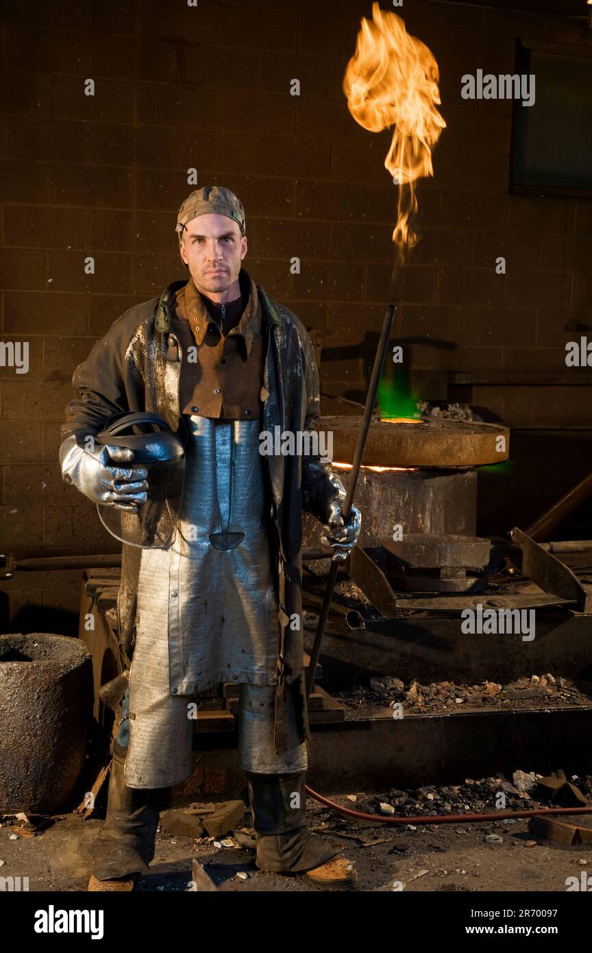 Ein Mann in einer Foudry-Jacke und Overalls, der eine Flammenröhre in einer Hand hält und eine Schutzmaske unter dem Arm hält, die in die Kamera einer Gießerei starrt. Stockfoto