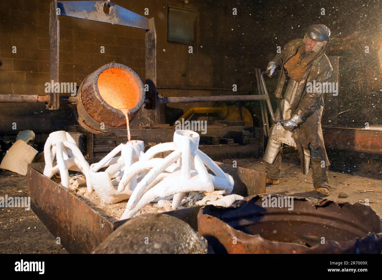 Ein Mann gießt orange geschmolzene Bronze in weiße Formen in einer Gießerei mit Hilfe eines Stützkrans. Stockfoto