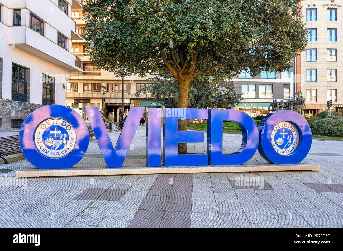 Oviedo, Asturias, Spanien: Eine Buchstabenskulptur mit dem Schriftzug „Oviedo“, umgeben von Fliesenboden in einem Stadtteil. Bäume, Stadtgebäude und Touristen Stockfoto