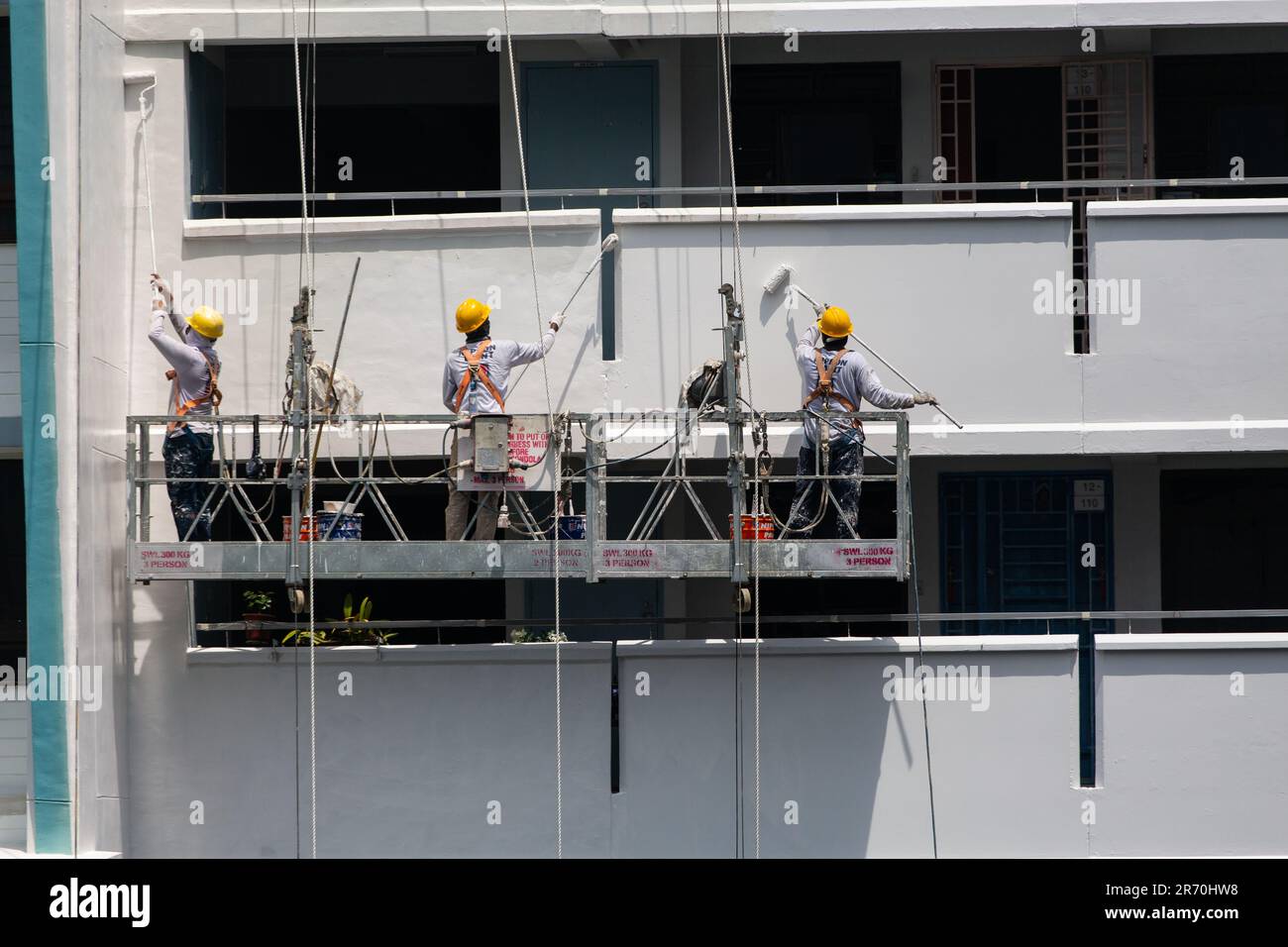 Arbeiter, die einen Industrieleiter benutzen, der in der Luft hängt und unter der heißen Sonne einen Gebäudeaußenbereich lackiert. Ein sehr risikoreicher Beruf. Stockfoto