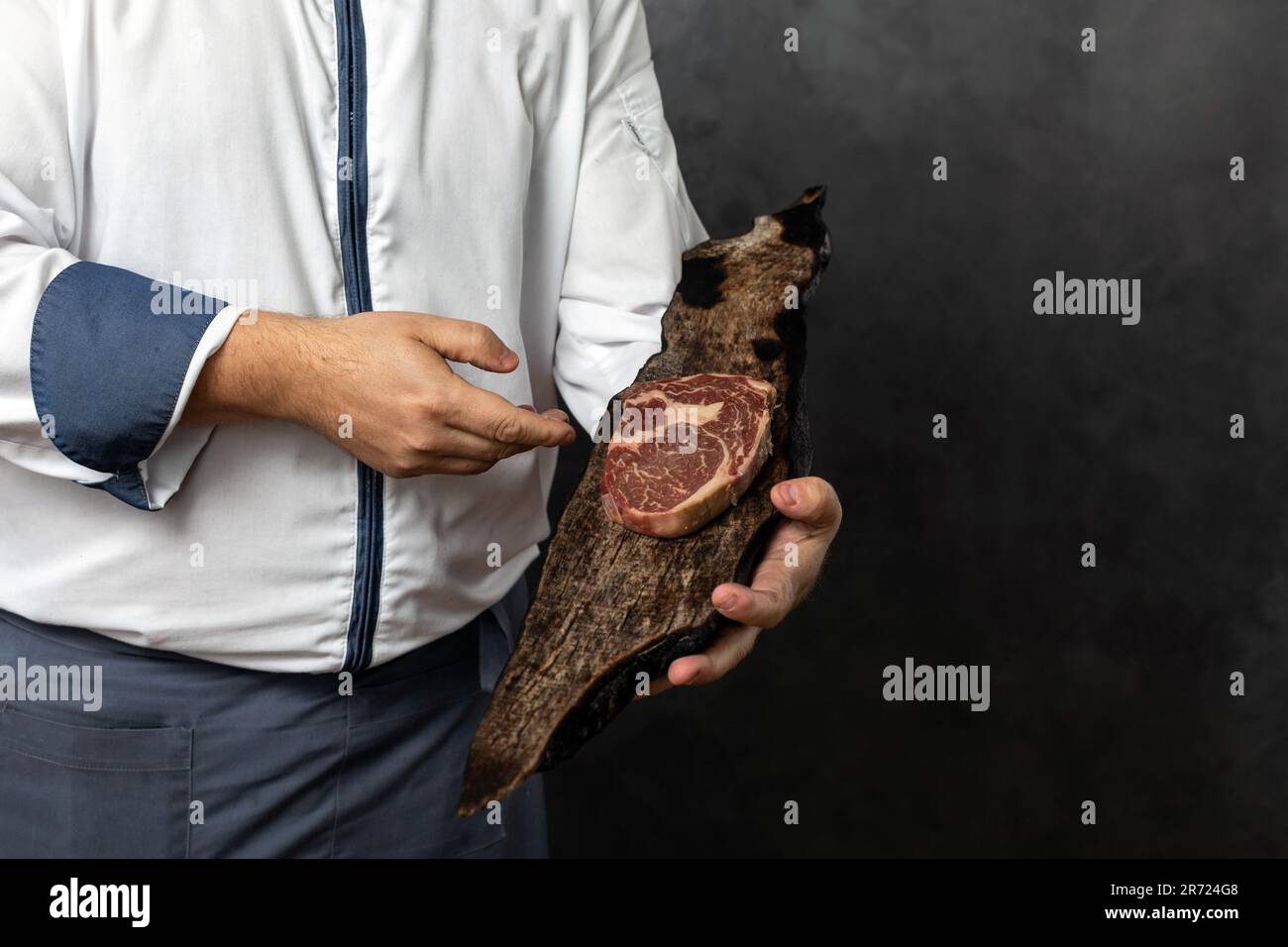 Ein anonymer männlicher in Uniform ernten und ein Stück frisches, köstliches Fleisch auf einem Holzbrett vor schwarzem Grunge-Hintergrund halten Stockfoto