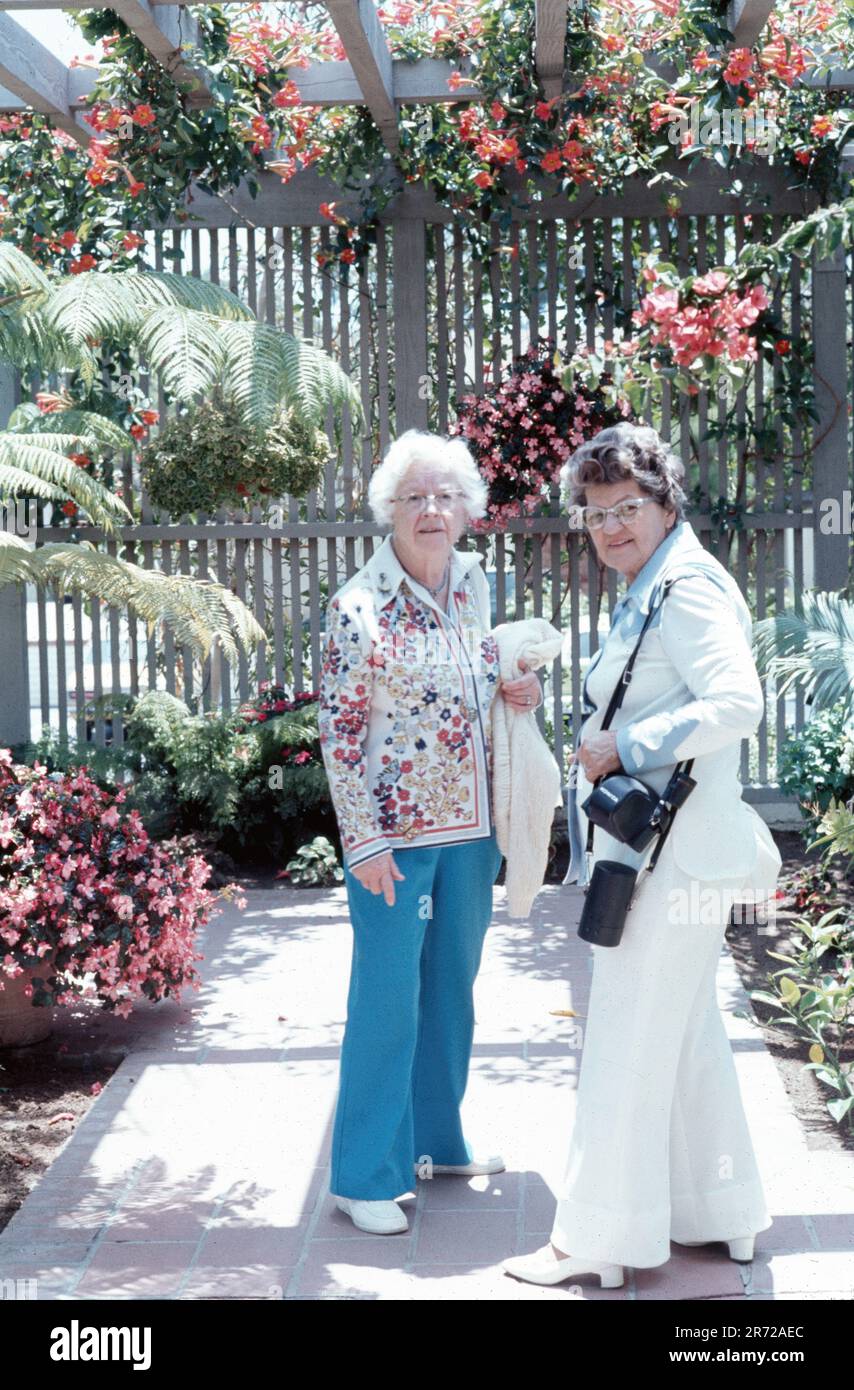 Zwei ältere Frauen stehen in einem Garten mit Blumen, 1975 US-Mode, 70s blauer Polyester, weißer Hosenanzug Stockfoto