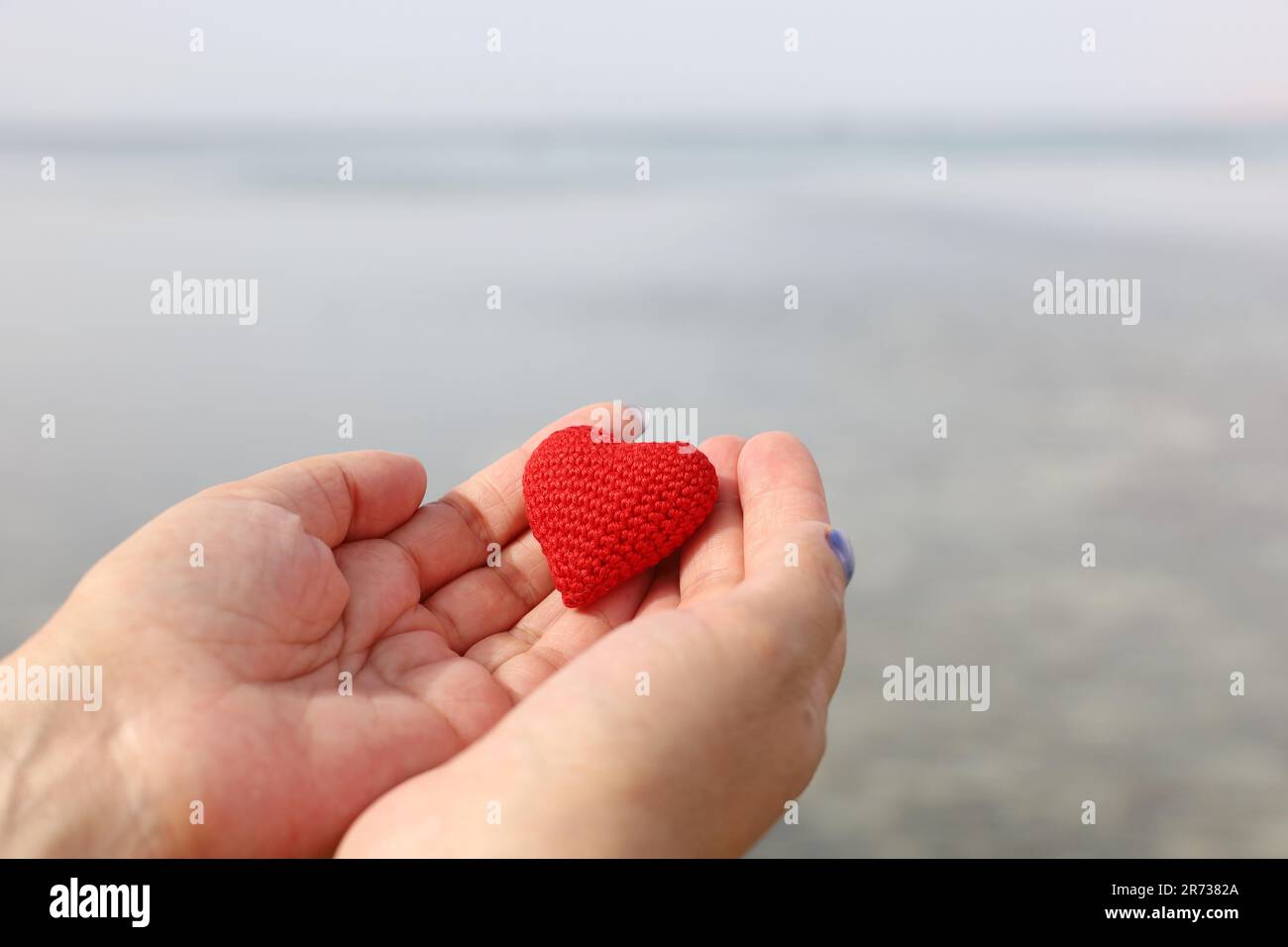 Seereise und Liebe am Sandstrand, rotes Strickherz in weiblichen Händen am Wasser. Valentinstag, Konzept einer romantischen Reise Stockfoto