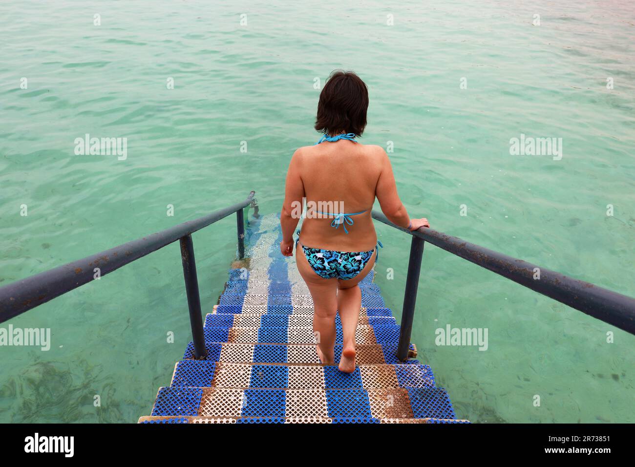 Eine Frau im Badeanzug geht die Leiter des Piers hinunter, um im Meer zu schwimmen. Strandurlaub und Schwimmen Stockfoto