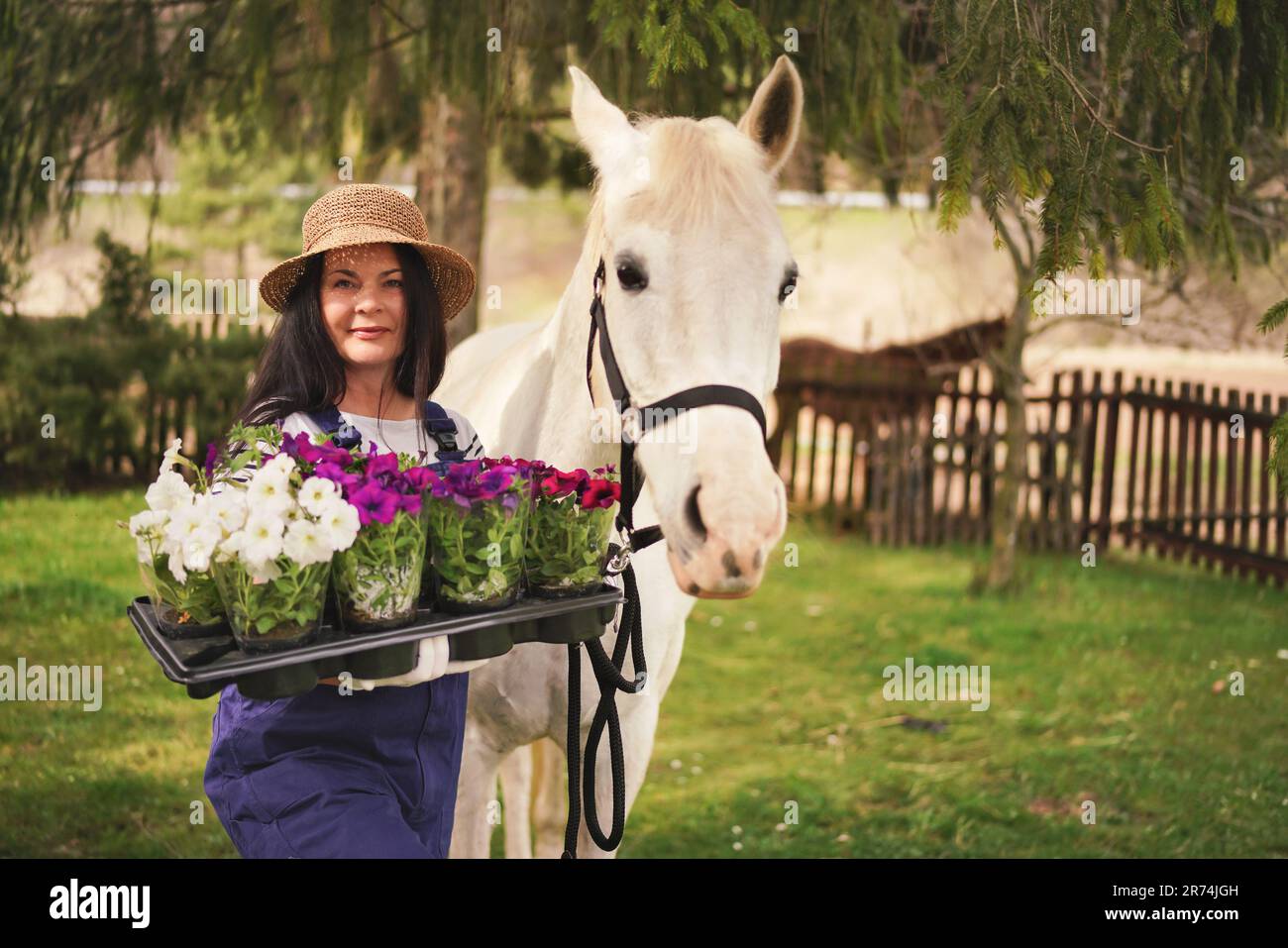 Frau in Arbeitskleidung, Handschuhen und Strohhut, die mit Blumen im Garten arbeitet, weißes Pferd in der Nähe, verschwommene grüne Bäume und Gartenhintergrund Stockfoto