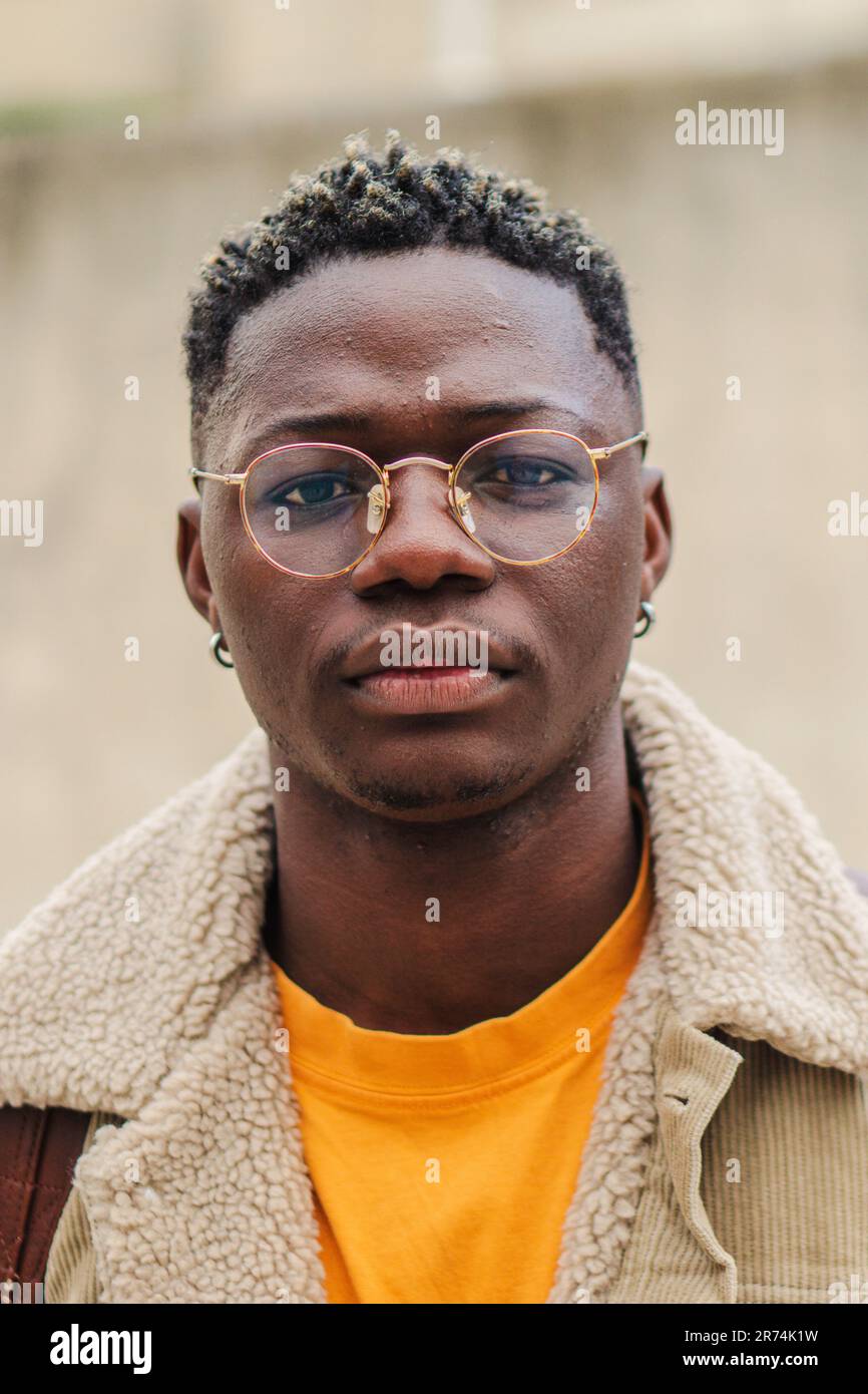 Vertikales Nahporträt eines klugen männlichen Teenagers mit Brillen auf dem Universitätscampus. Junger afroamerikanischer Student, der ernsthaft vor die Kamera schaut. Selbstbewusster Kerl mit nachdenklicher Einstellung. Hochwertiges Foto Stockfoto