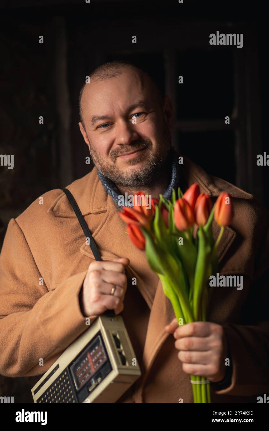 Ein altmodischer Mann mittleren Alters, der lächelt und in die Kamera schaut, mit einem Blumenstrauß und einem Retro-Radio auf dunklem Hintergrund. Stockfoto