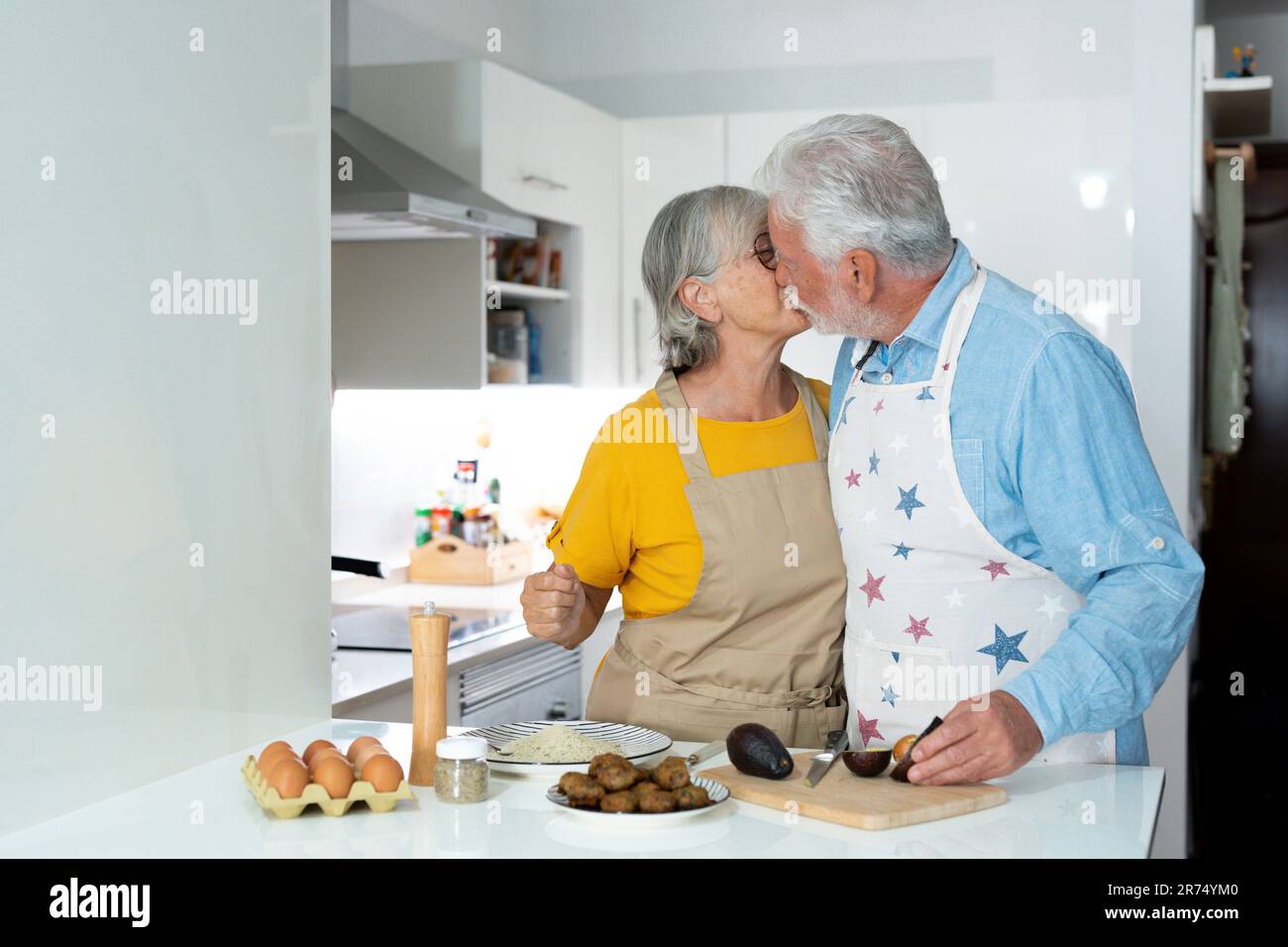 Glücklicher, glücklicher älterer Mann und Frau, die sich zusammen küssen, Bio-Salat zusammen machen, reden, lachen. Ein altes Paar, das Spaß in der Küche hat, das Abendessen zubereitet Stockfoto