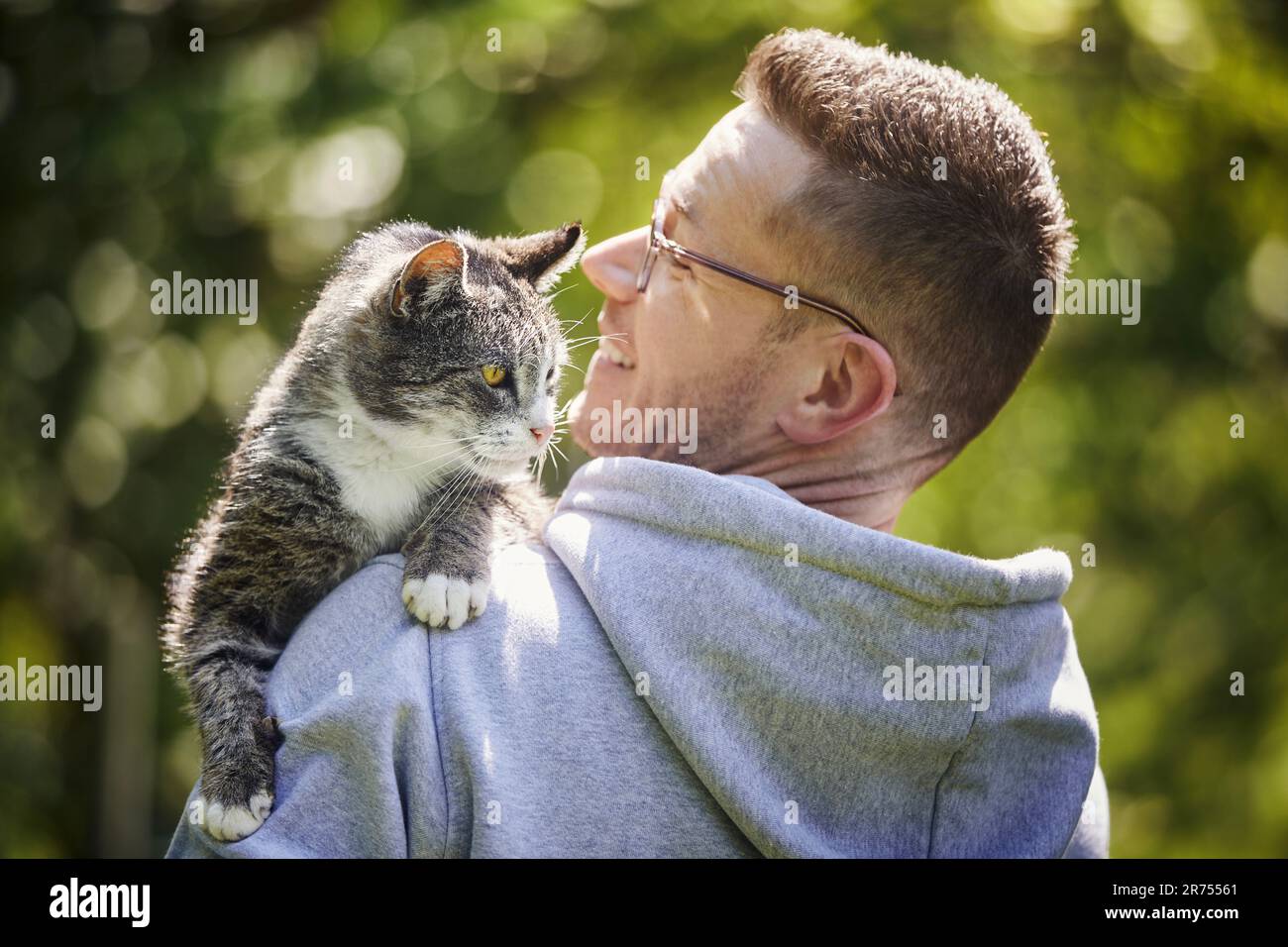 Lächelnder Mann, der seine neugierige Katze auf der Schulter trägt. Haustierbesitzer mit alter Tabby-Katze im Garten an sonnigen Tagen. Stockfoto