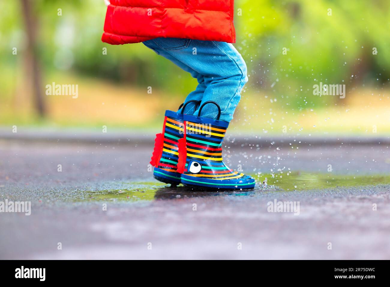 Die Füße eines Kindes in bunten Gummistiefeln springen über eine regnerische Pfütze in einem Park Stockfoto