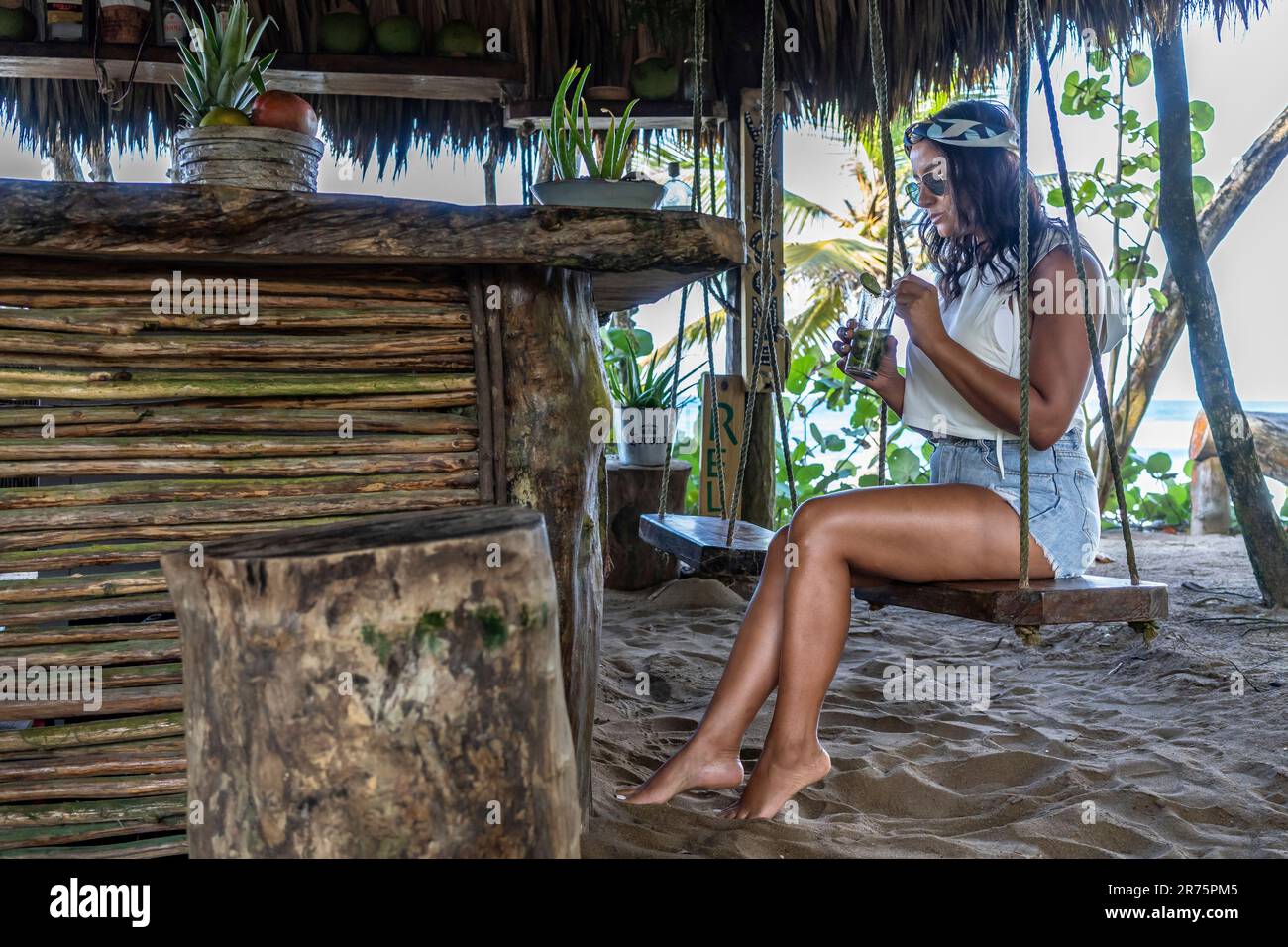 Nordamerika, Karibik, Großantillen, Hispaniola, Dominikanische Republik, North Coast, Puerto Plata Province, Cabarete, Natura Cabana, attraktive Latina sitzt auf Einer Schaukel an der Beach Bar und trinkt einen Drink Stockfoto