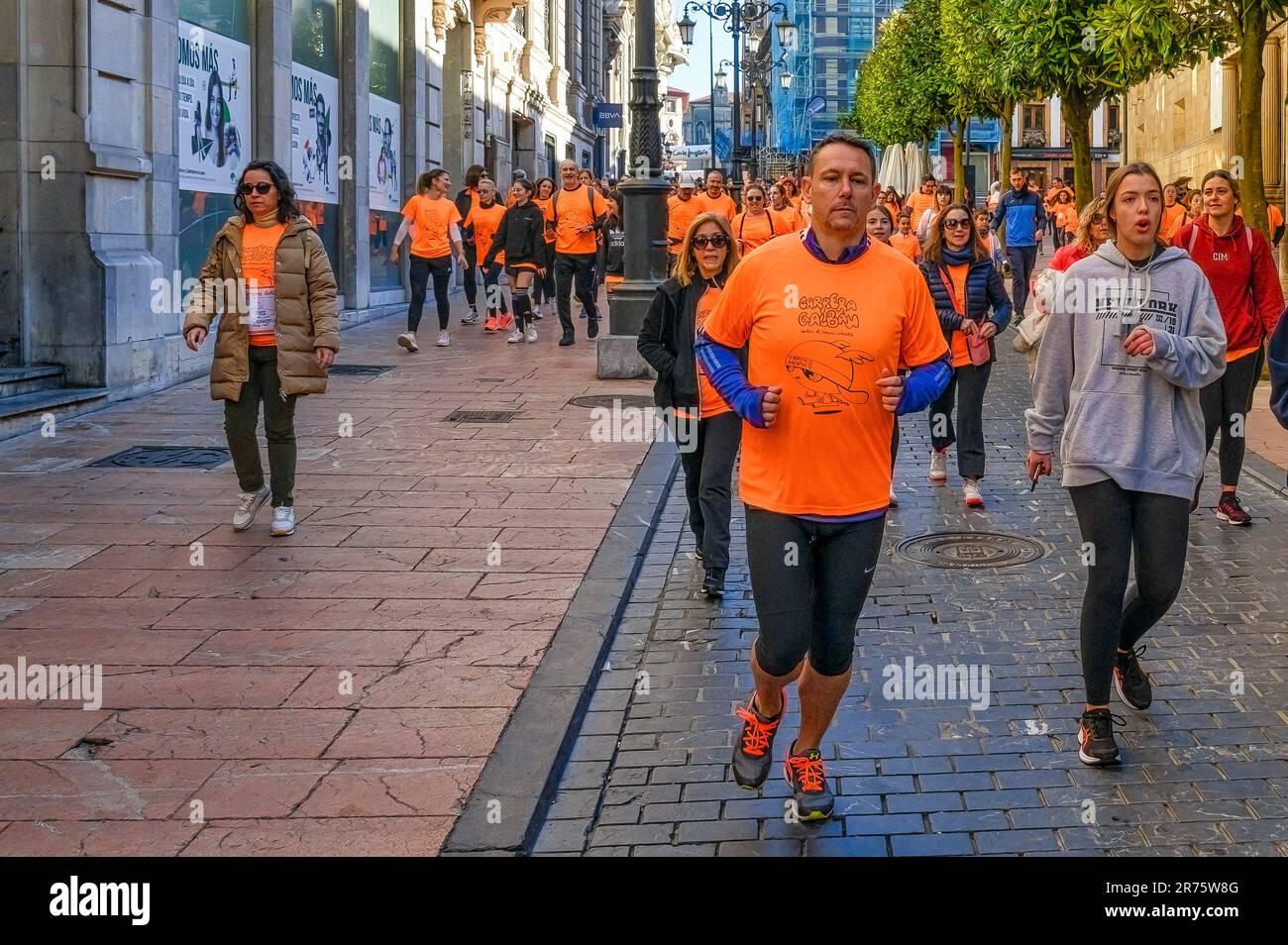 Oviedo, Asturien, Spanien - 12. Februar 2023: Menschen joggen auf einer gefliesten Stadtstraße und einem Bürgersteig, viele tragen orangefarbene T-Shirts. Gebäude und Baum Stockfoto